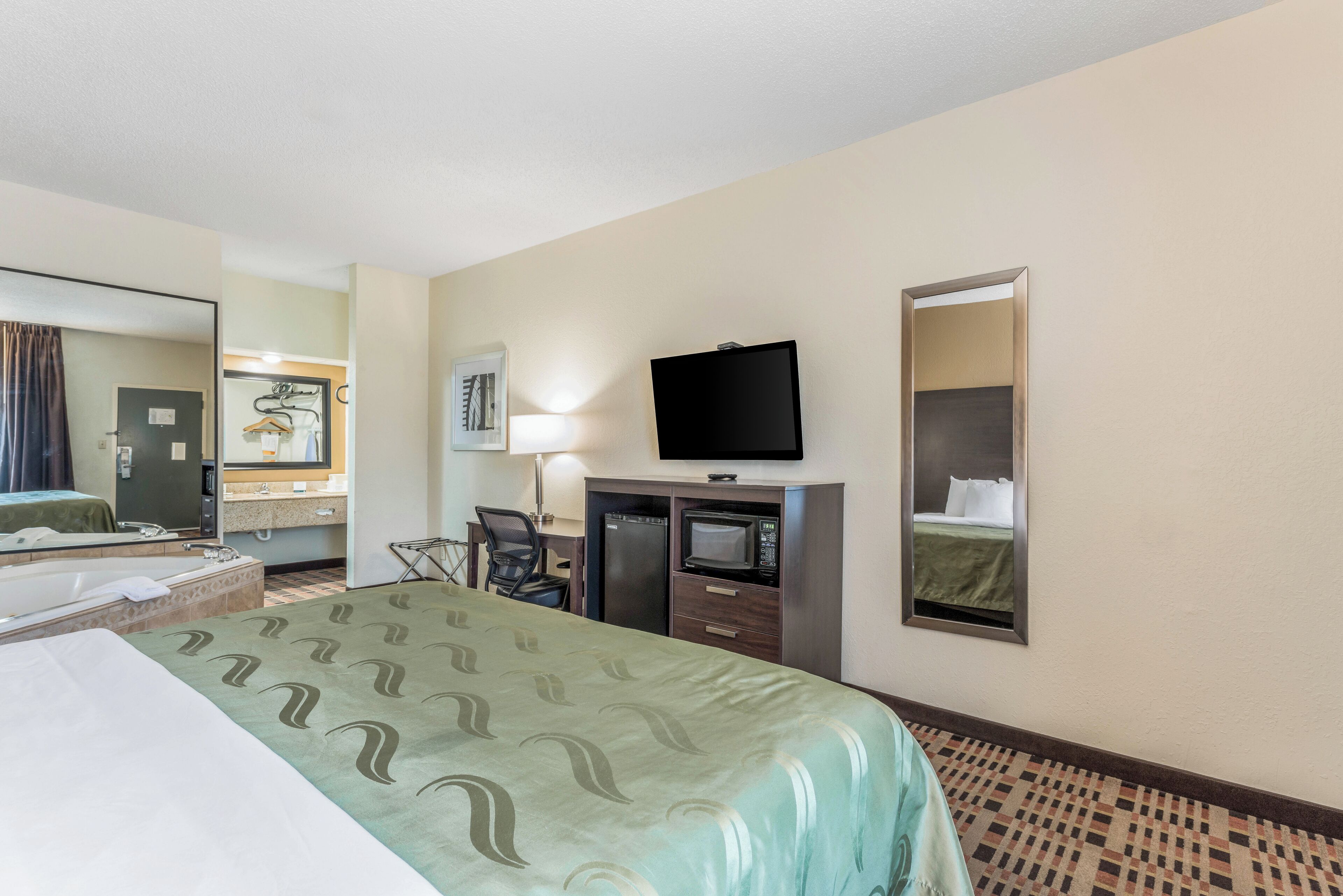 A hotel room with a bed, a desk with a chair, a television, a microwave, and a mirror.