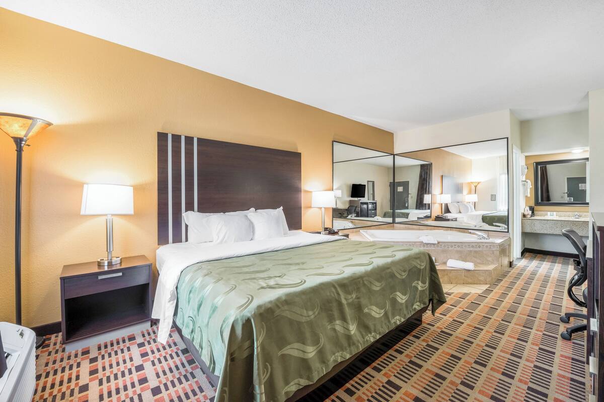 Une chambre d’hôtel comprenant un lit, une table de chevet, une lampe, un miroir et une salle de bain avec une baignoire et un lavabo.