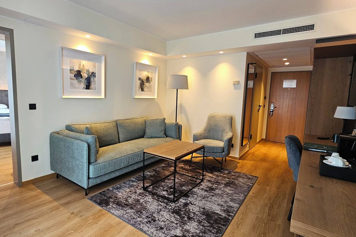 A modern hotel room with a grey sofa, a small wooden table, a floor lamp, and a rug.