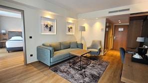 Ein modernes Hotelzimmer mit grauem Sofa, einem kleinen Holztisch, einer Stehlampe und einem Teppich.