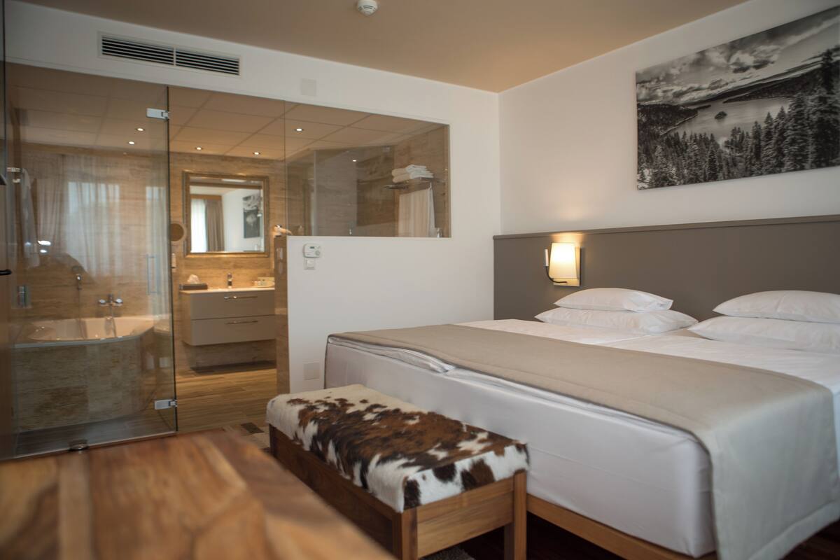 A hotel room with a large bed, a wooden bench, a glass shower enclosure, a bathroom with a mirror, and a painting on the wall.