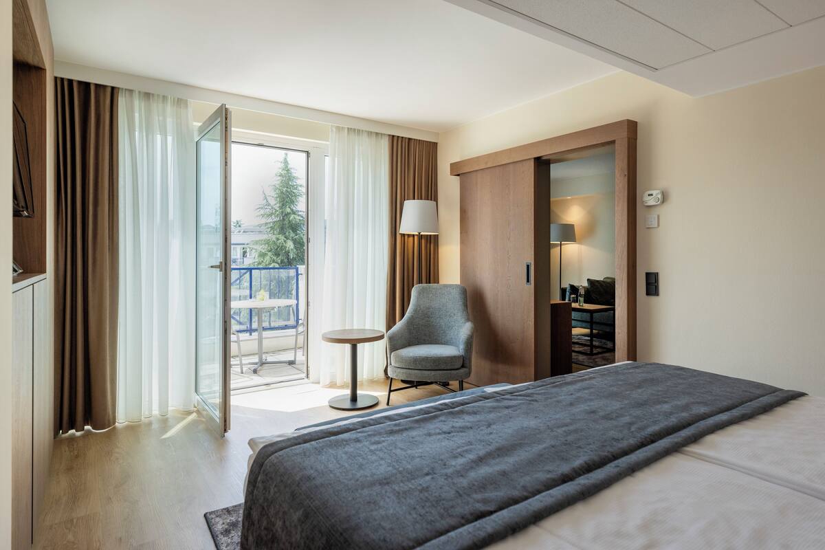 A hotel room with a bed, a chair, a small table, and a sliding door leading to an outdoor area.