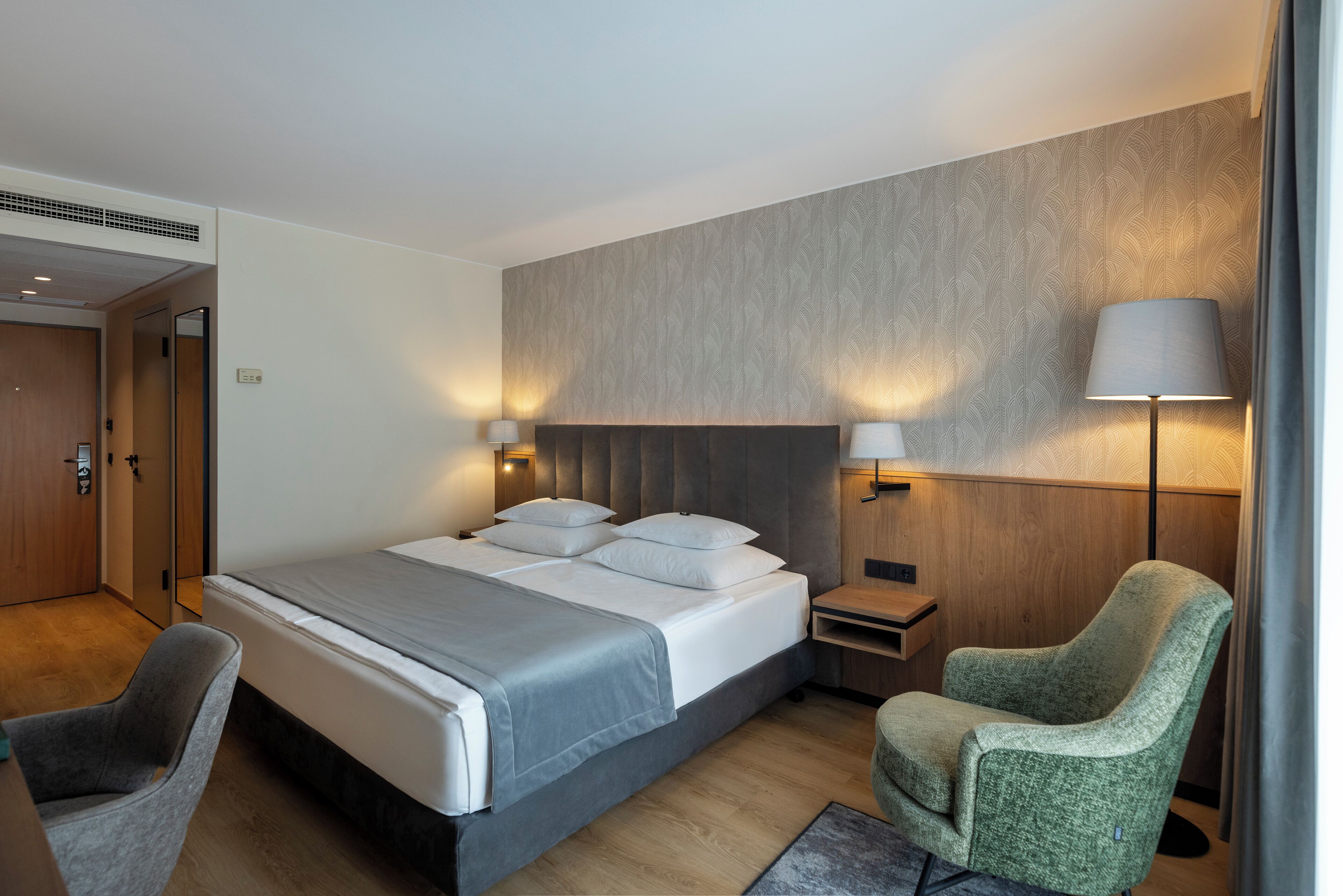 A modern hotel room with a large bed, two bedside lamps, a green armchair, and a wooden headboard.