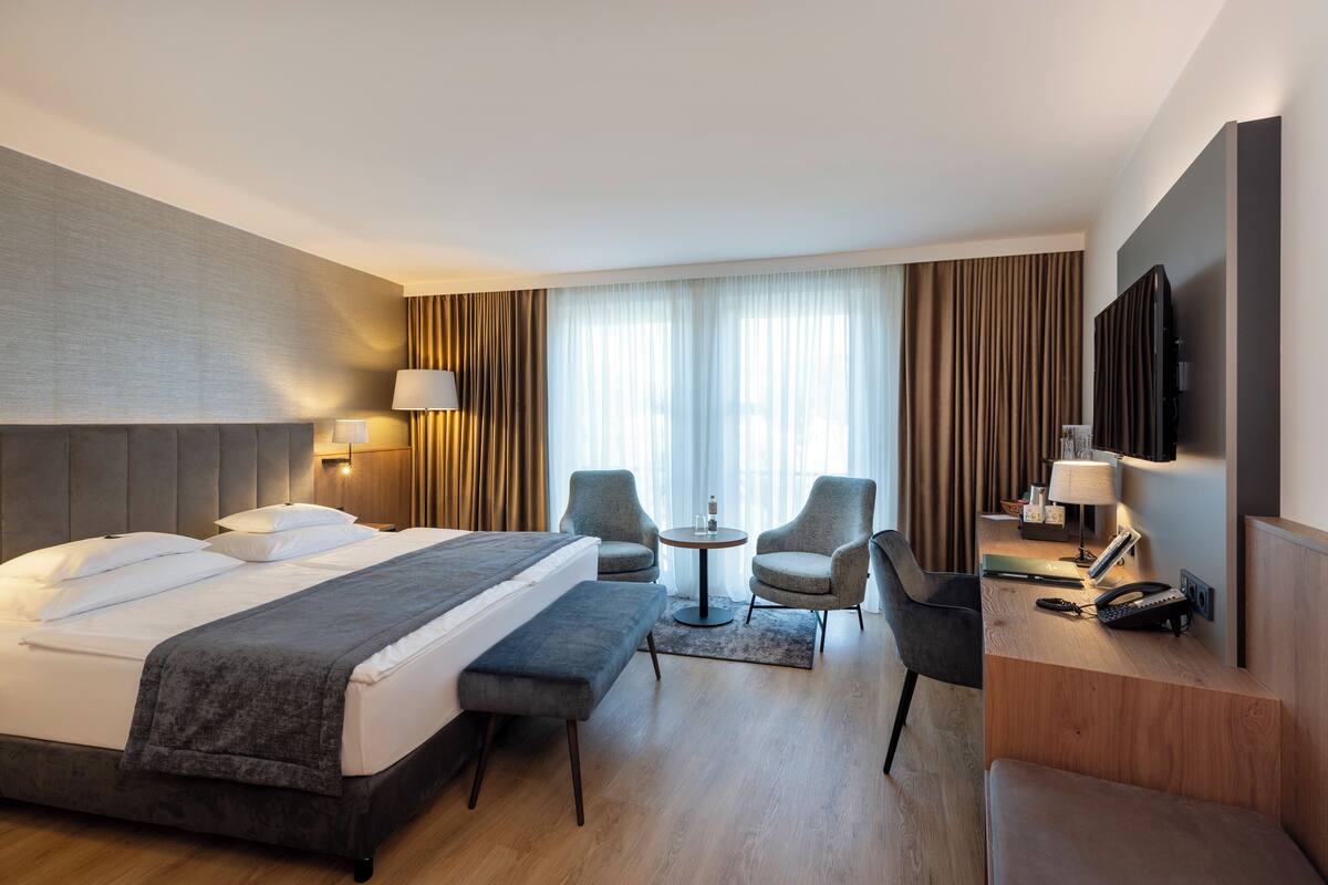 A hotel room with a large bed, two armchairs, a small table, a desk with a telephone, and a TV mounted on the wall.
