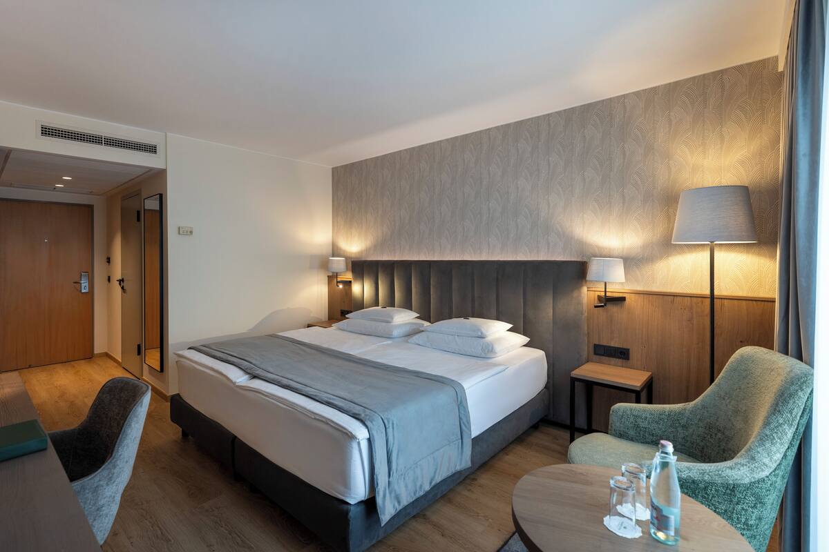 A hotel room with a bed, bedside tables, a chair, and a small table with a lamp and glasses.