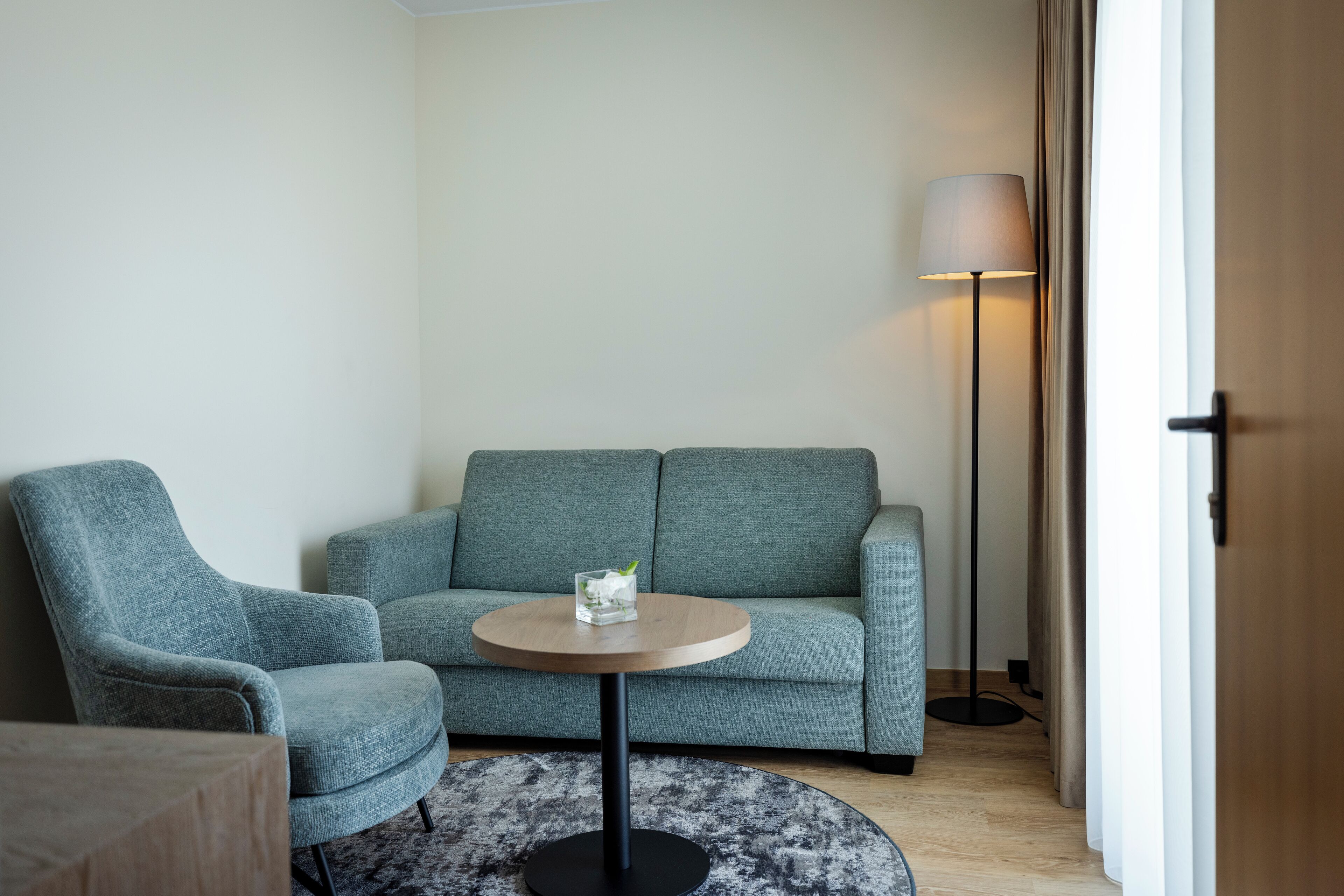 Une pièce avec un canapé gris, une table ronde en bois sur laquelle se trouve un objet en verre, un lampadaire et une porte en bois.