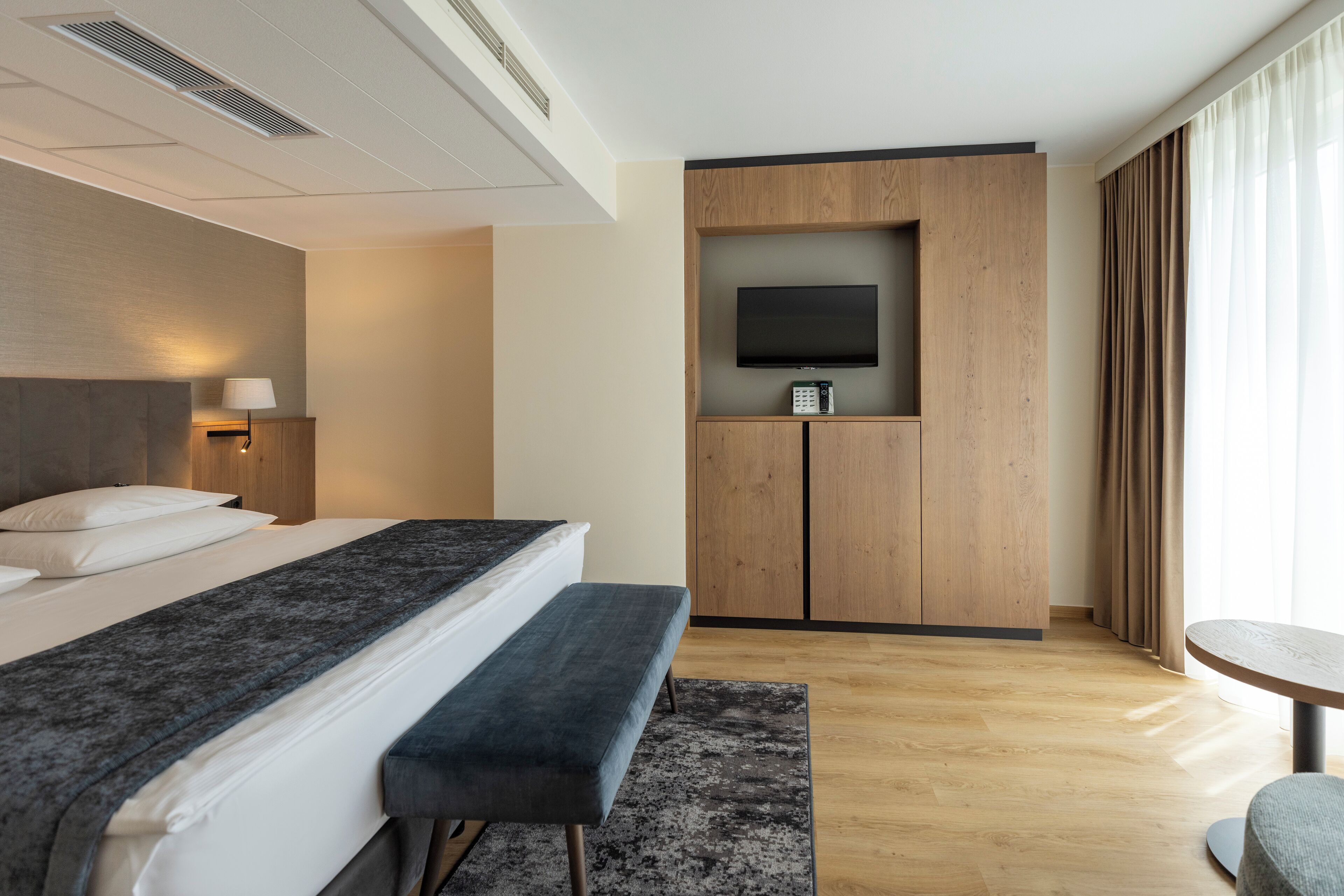 Quarto de hotel moderno com uma cama grande, um banco, uma mesa pequena, uma televisão presa a um móvel de madeira e uma janela com cortinas.