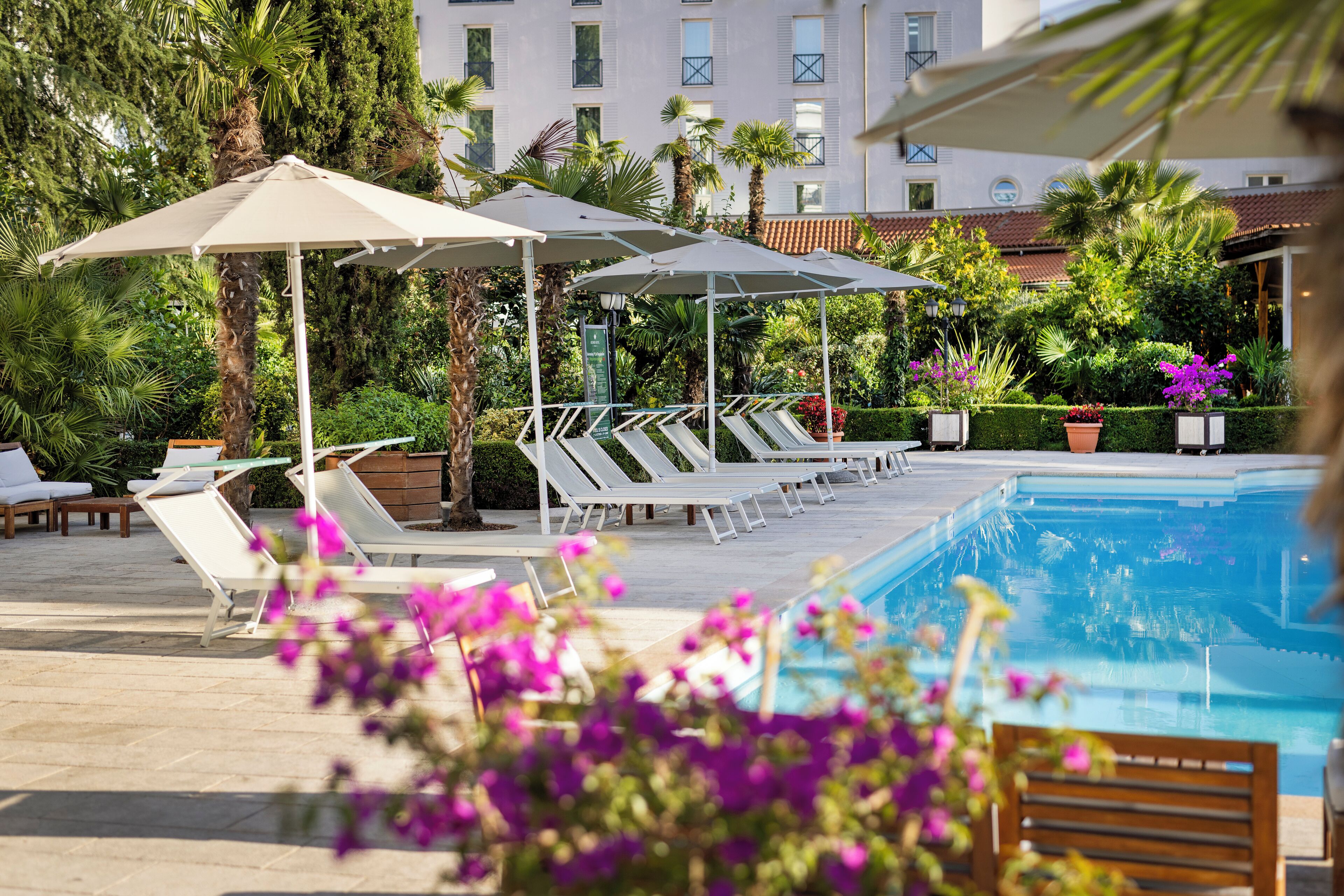 Un área de piscina con sillones de playa y sombrillas, rodeada de follaje y un edificio al fondo.