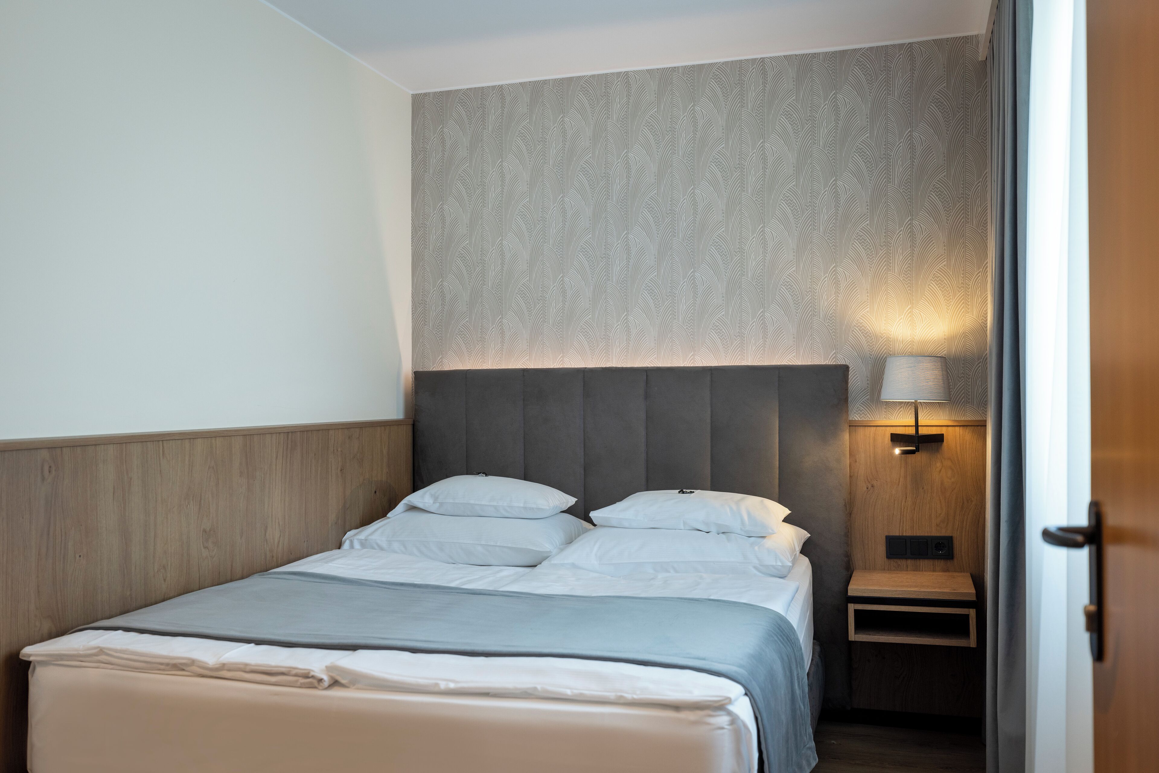 A neatly made bed with a grey bedspread and white pillows, a wooden headboard, and a bedside table with a lamp.
