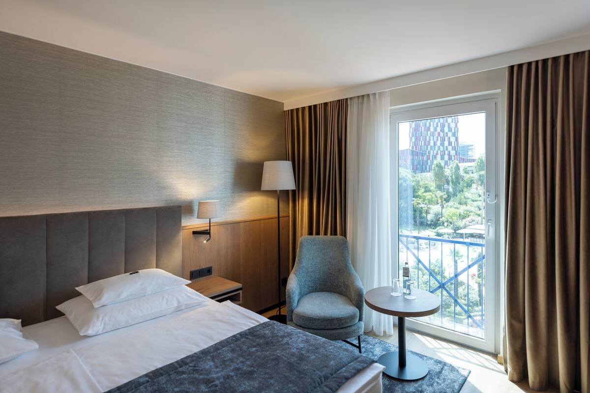 A hotel room with a bed, a chair, a small table, a lamp, and a view of the city through large windows.