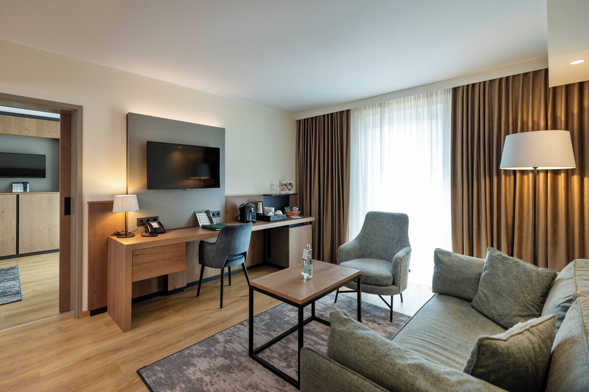 A modern hotel room with a wooden desk, a comfortable sofa, a flat-screen TV, and a large window with curtains.