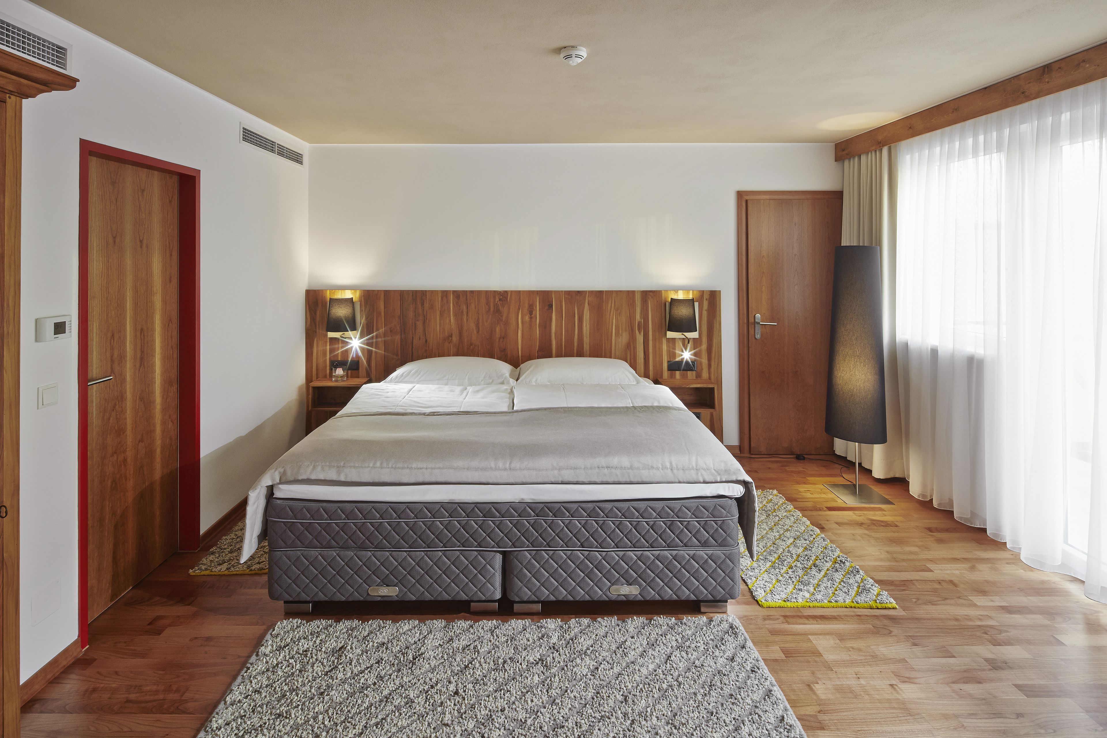 A modern hotel room with a large bed, wooden headboard, bedside lamps, a flat-screen TV, and a view of the outdoors through sheer curtains.