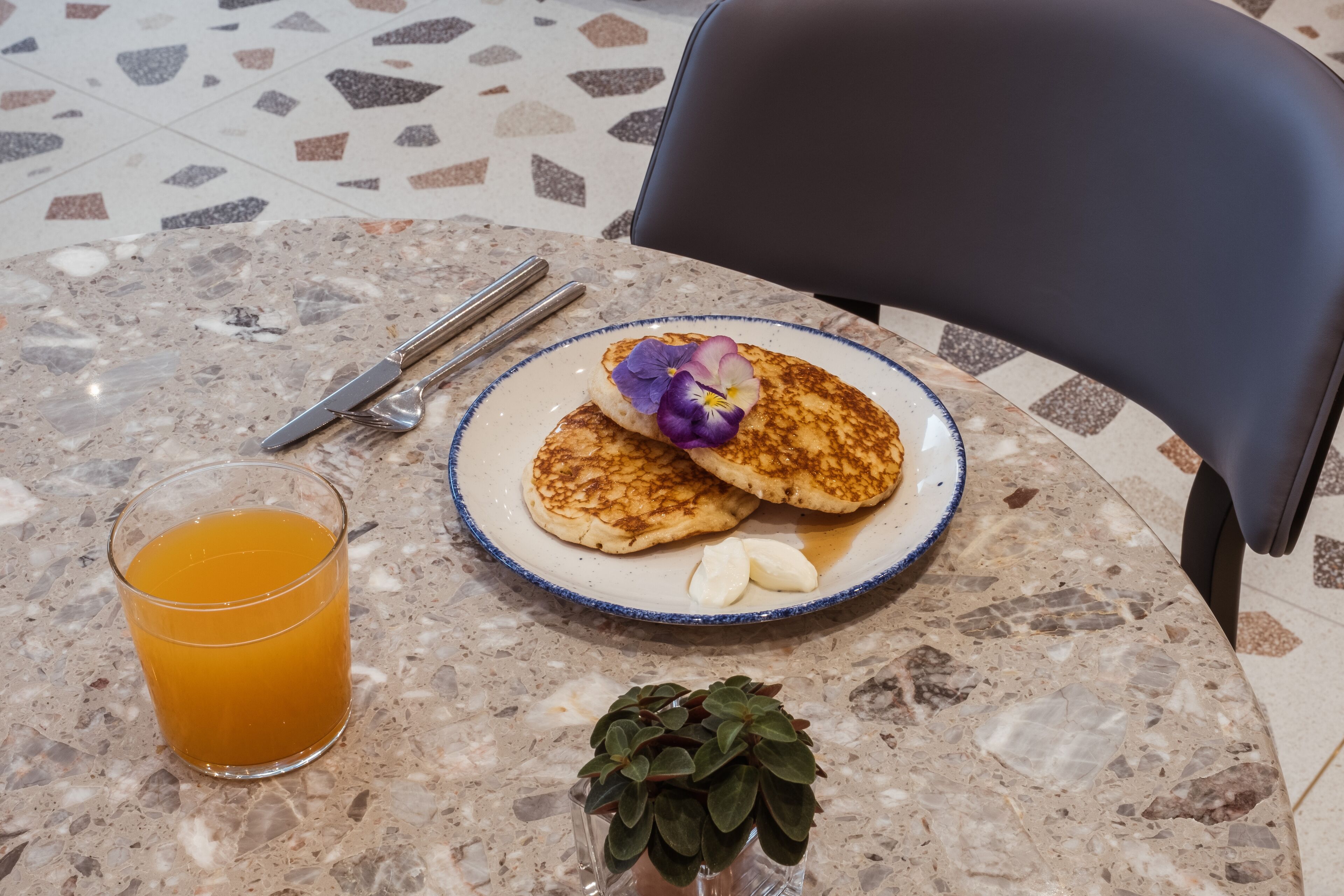 A breakfast setting with pancakes, a glass of orange juice, and a small plant on a marble table.