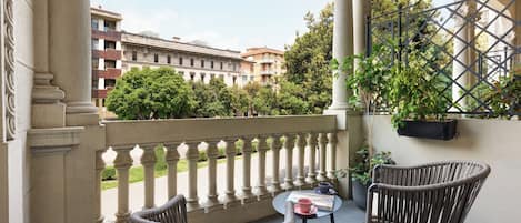 A balcony with wicker chairs and a small table, overlooking a courtyard with trees and buildings.