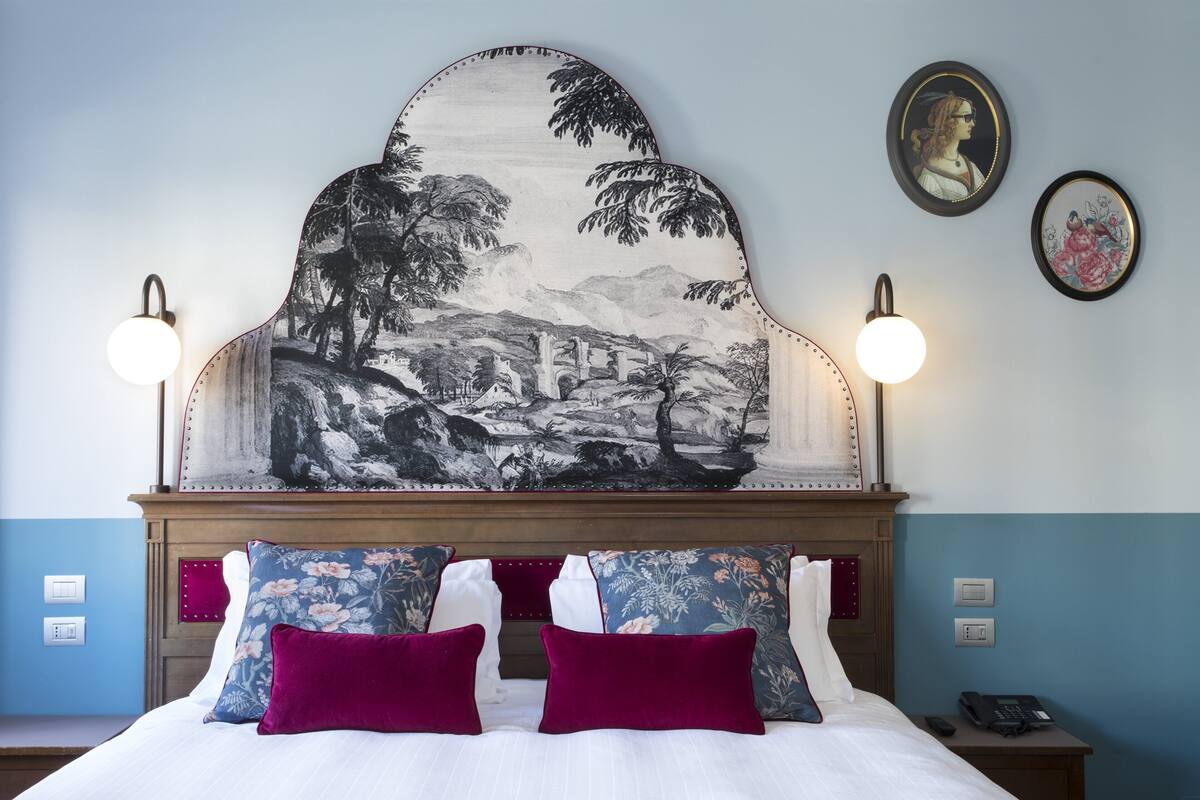 A hotel room with a bed, a headboard with a landscape painting, two wall sconces, and a nightstand with a phone.