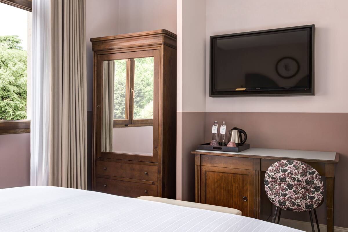 A hotel room with a bed, a wooden wardrobe, a small desk with a chair, a flat-screen TV, and a window with curtains.