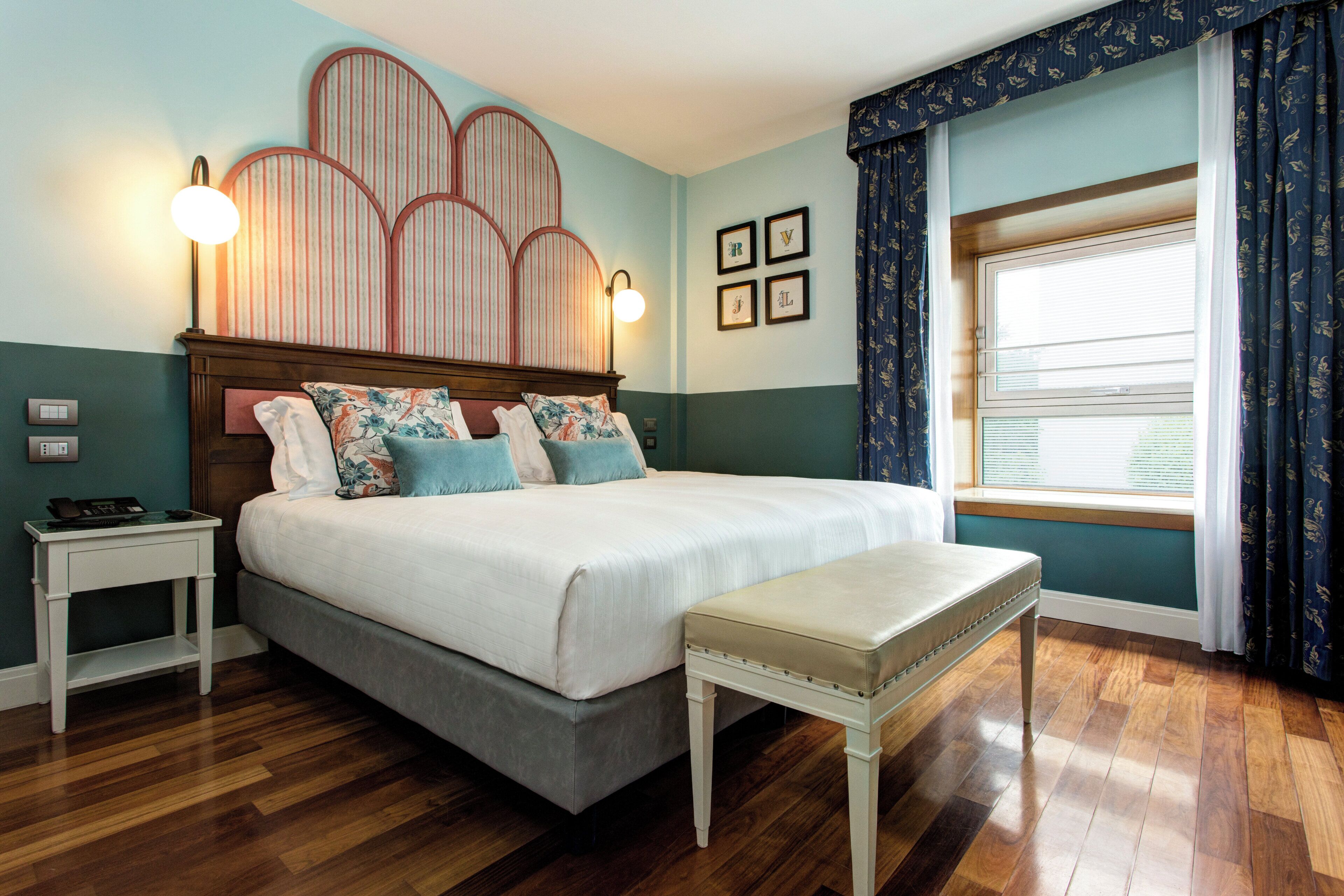 A bedroom with a bed, bedside tables, a bench, a window with curtains, and wall-mounted lights.