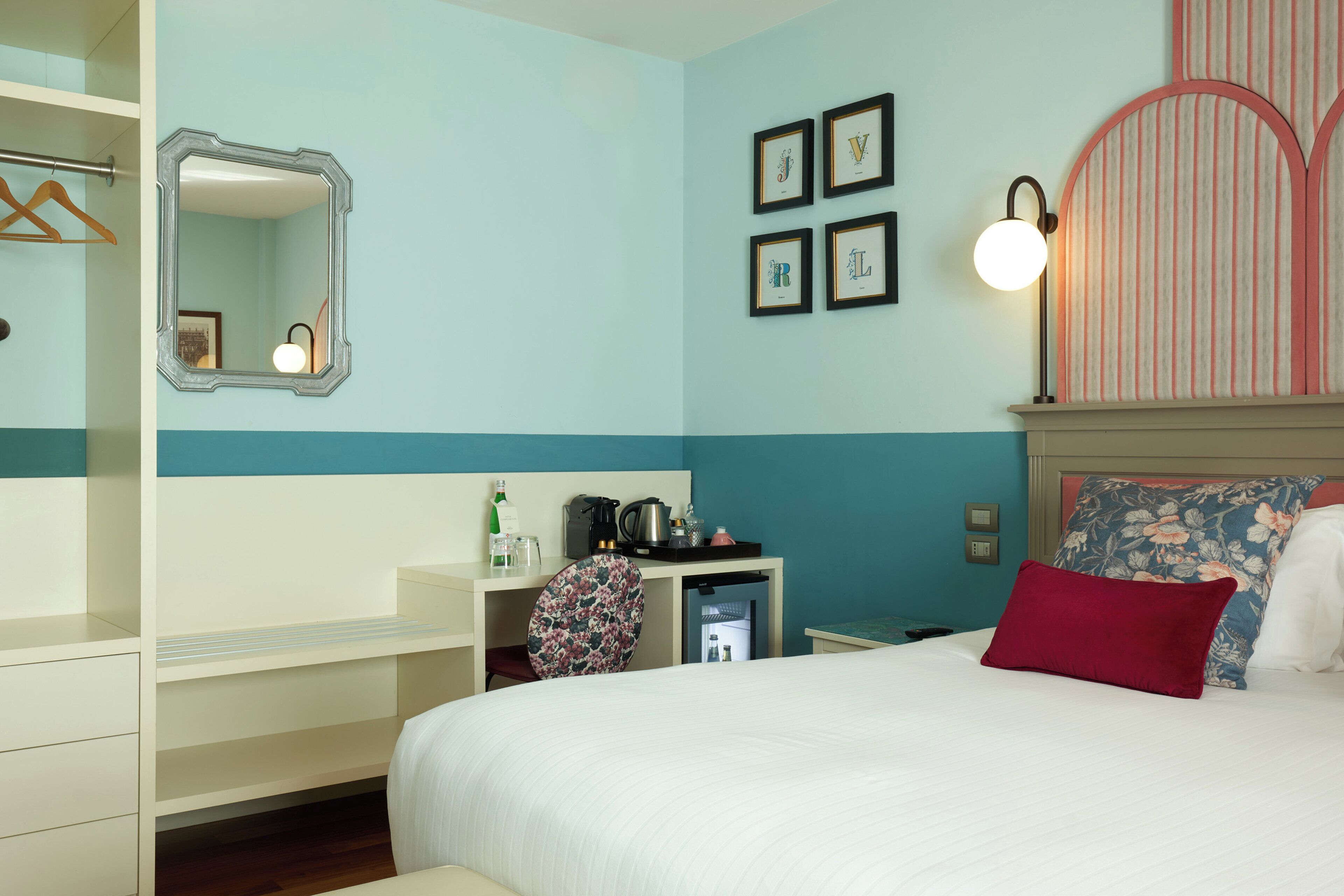 Habitación de hotel con cama, escritorio con cafetera, espejo y cuadros en la pared.