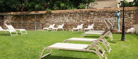 A patio area with several white lounge chairs arranged on a grassy lawn, a stone wall in the background, and a statue of a person holding a camera.