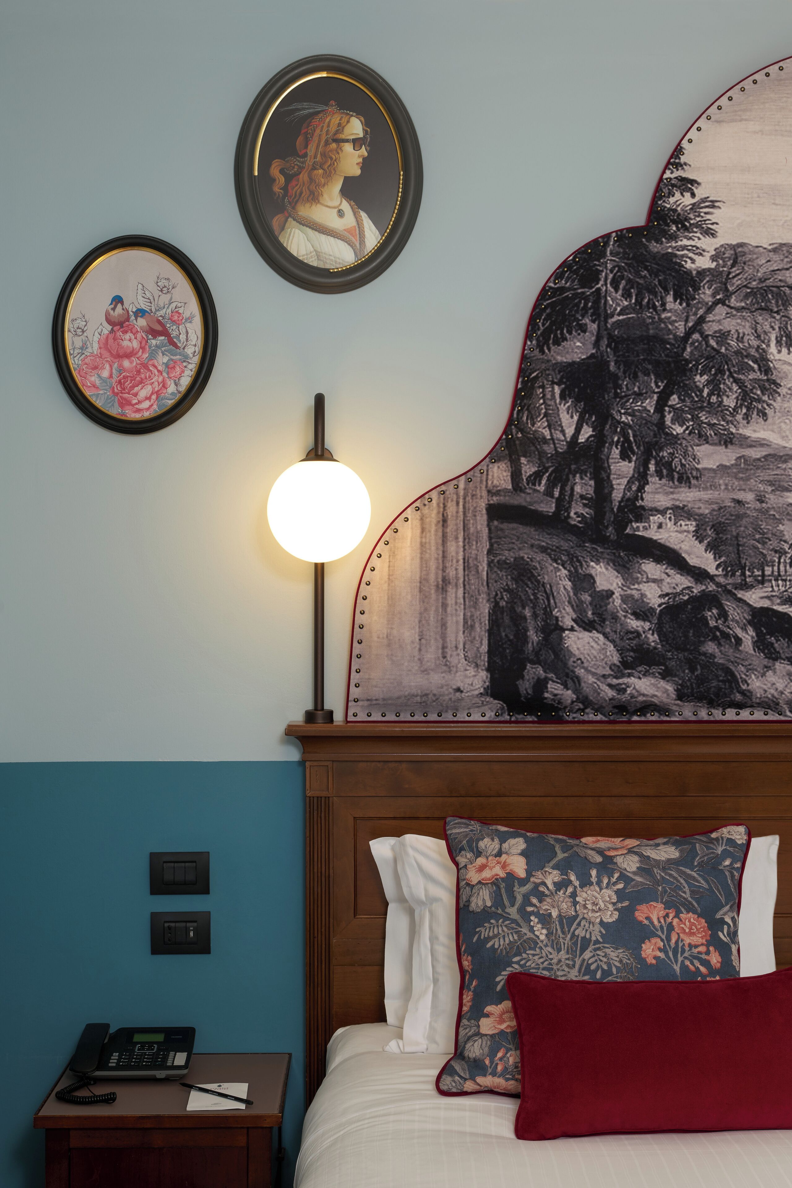 A hotel room with a bed, a nightstand with a phone, and wall art including a landscape painting and framed pictures.