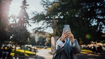 Pessoa segurando um smartphone, possivelmente tirando uma foto, em um parque com árvores e um caminho.