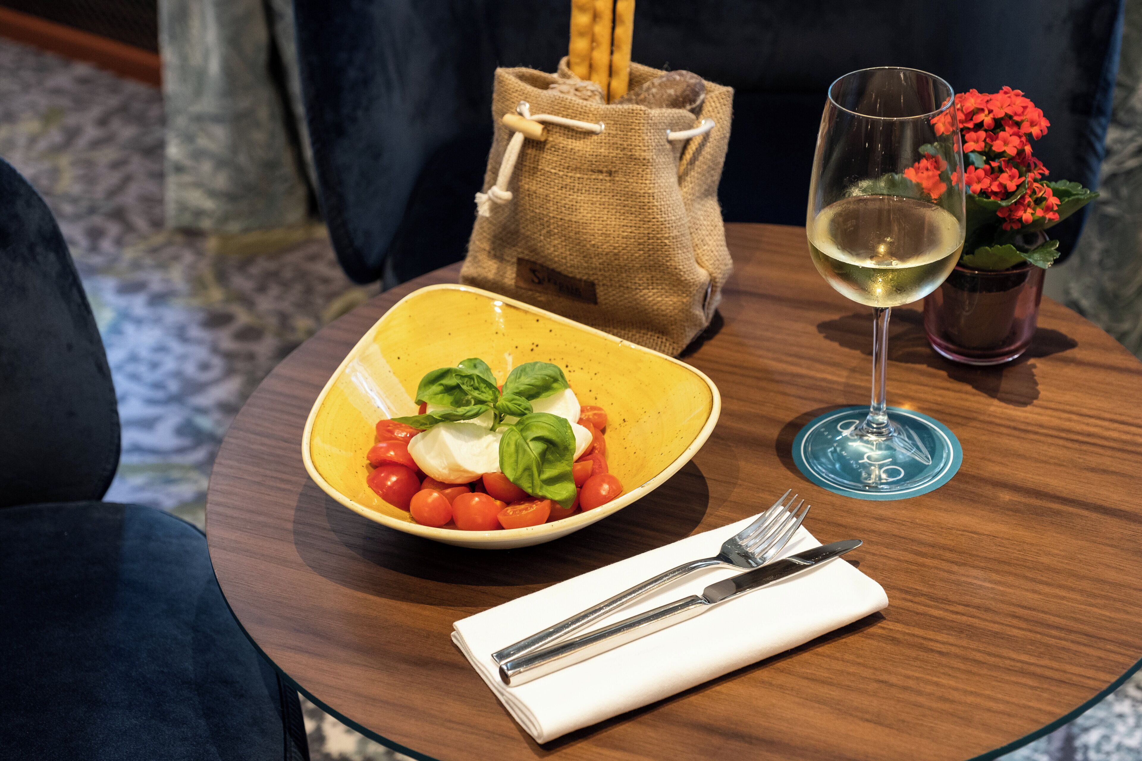 A table with a bowl of salad, a glass of white wine, a napkin with cutlery, and a small potted plant.