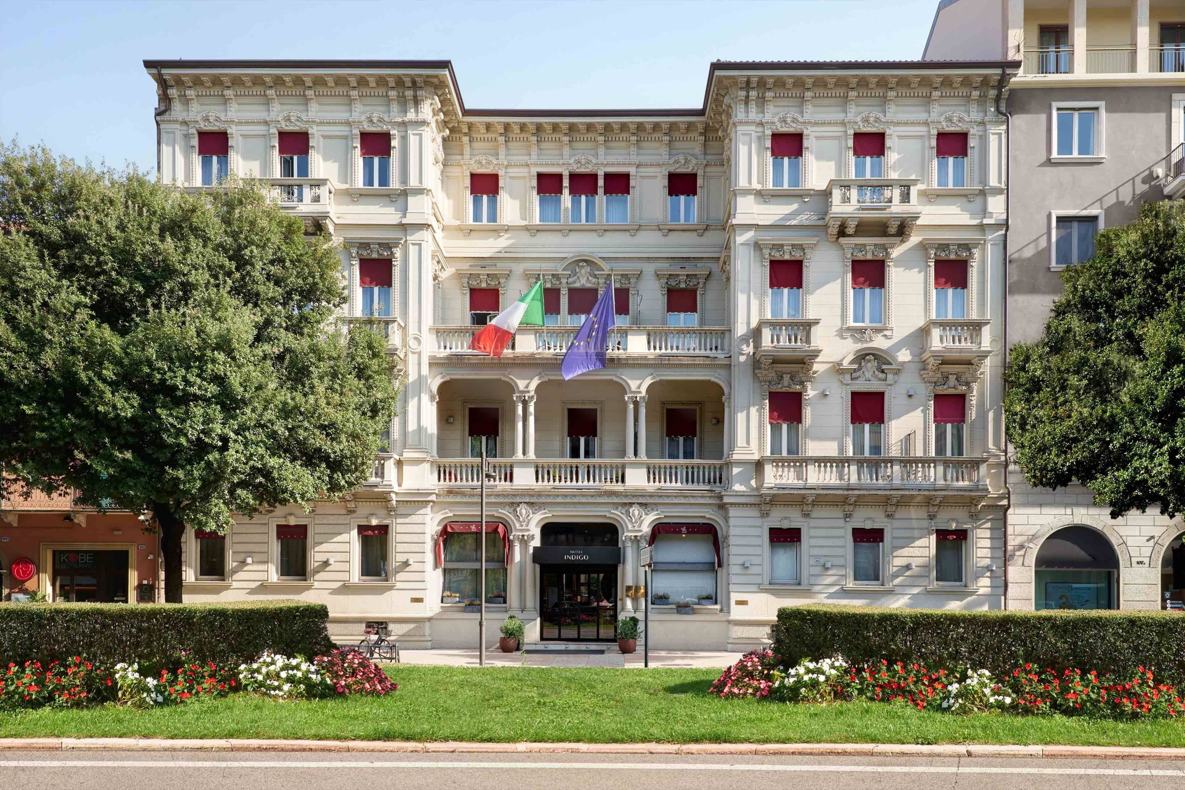 Un edificio de varios pisos con estilo arquitectónico clásico, que cuenta con una entrada central con toldo y las banderas de Italia y la Unión Europea.
