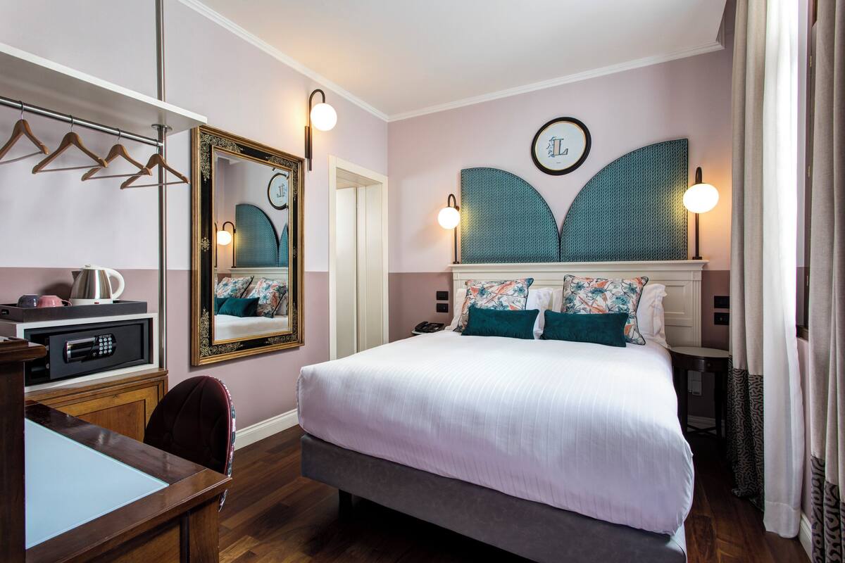 A hotel room with a large bed, a desk with a computer, a mirror, and wall-mounted lights.