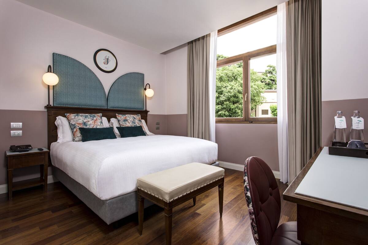 A hotel room with a large bed, a desk, a chair, a small table, and a window with curtains.
