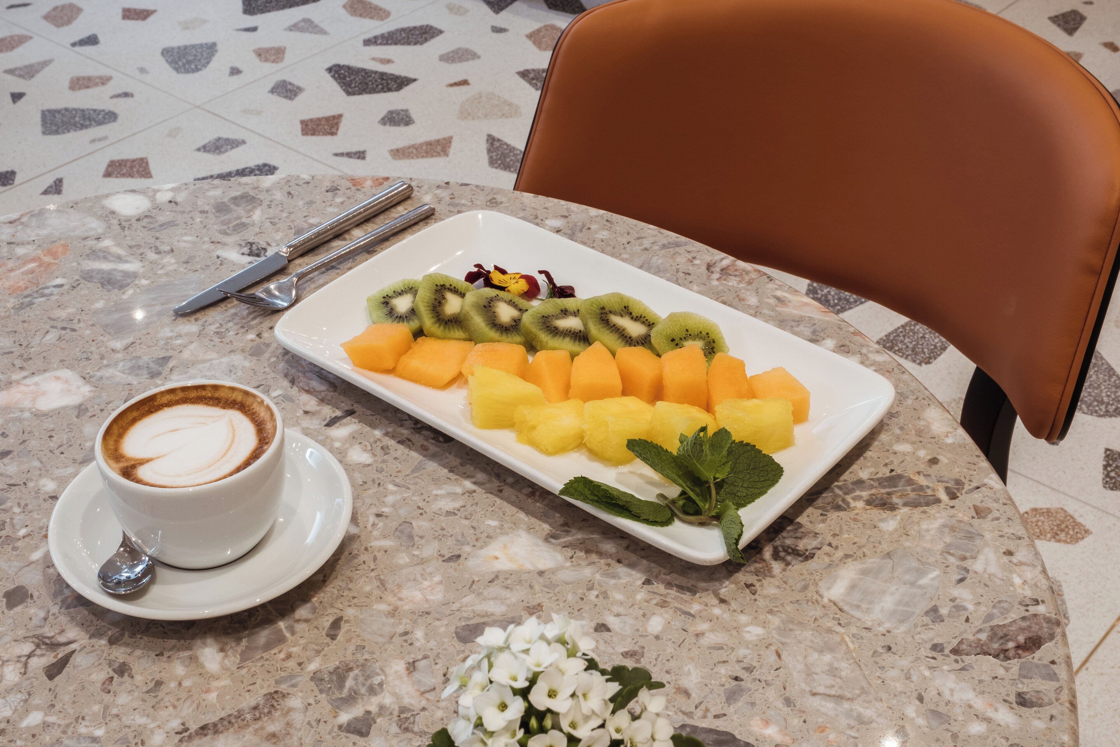 A marble table with a white plate of fruit, a cup of coffee, and a pair of cutlery.