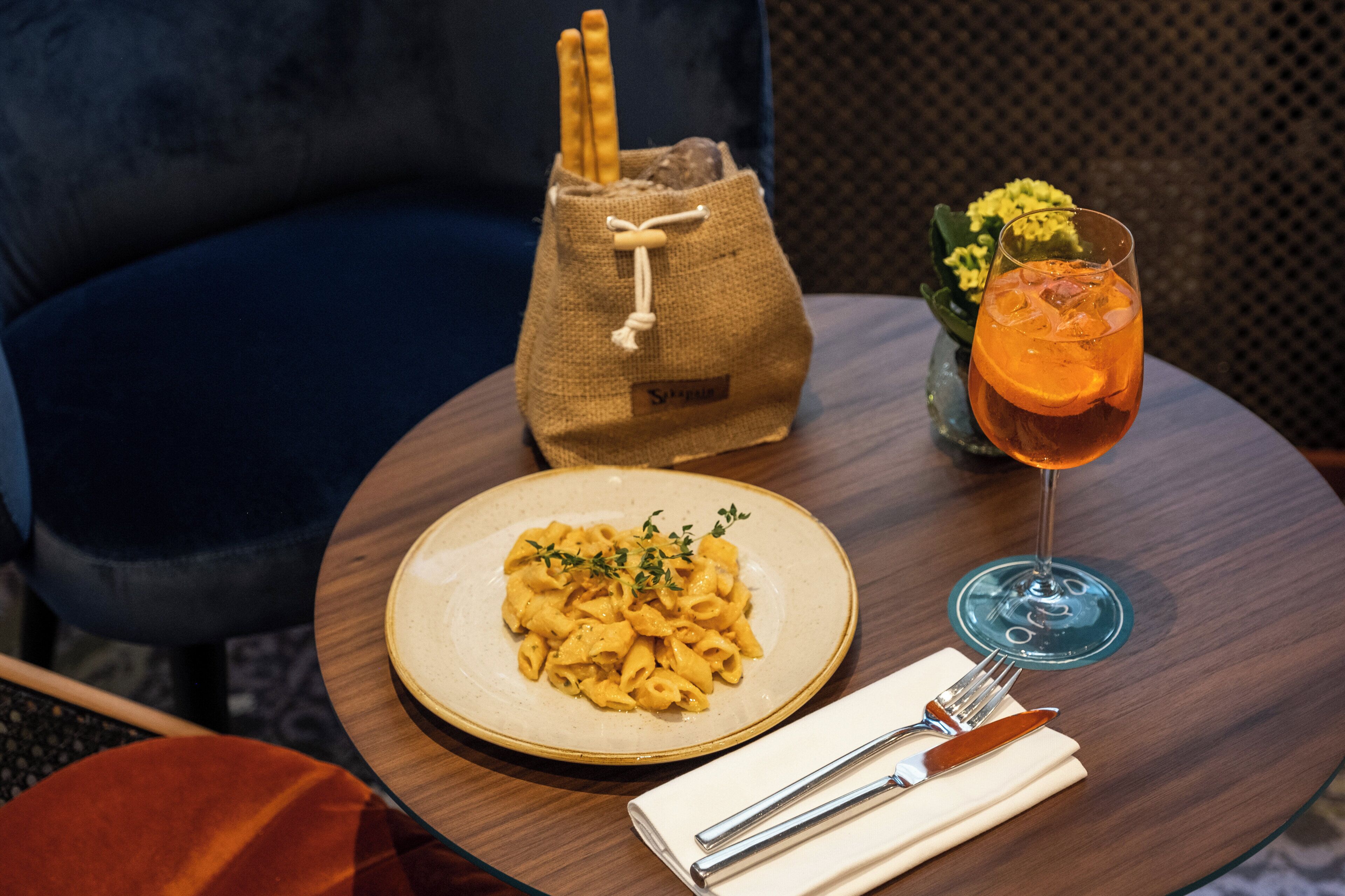 A table with a plate of pasta, a glass of orange drink, a small vase with flowers, and a bag.