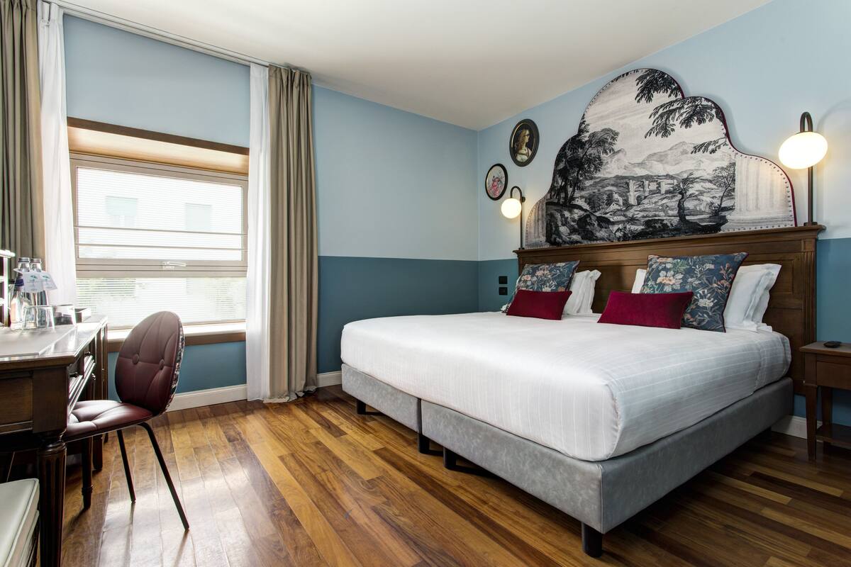 A bedroom with a large bed, a desk with a chair, a window with curtains, and a wall with framed artwork.