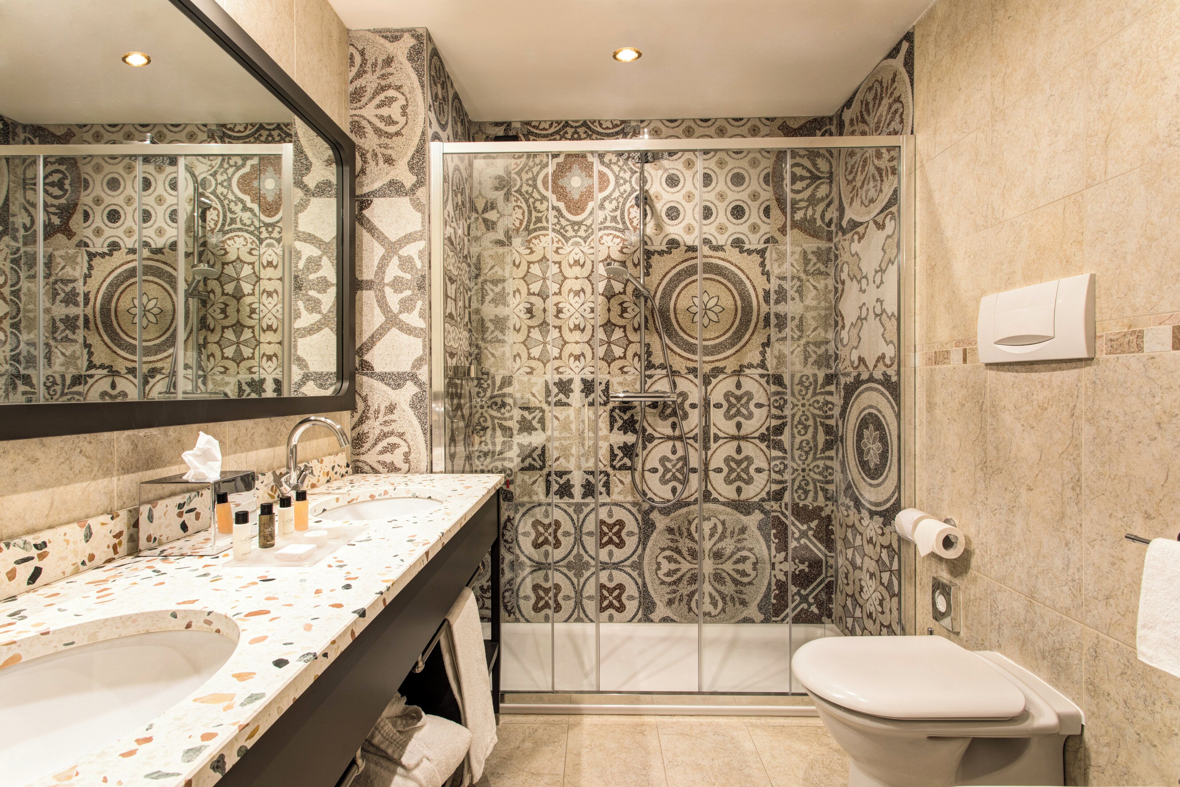 Une salle de bain avec un grand miroir, un lavabo avec des articles de toilette, une douche aux carreaux décorés et des toilettes.