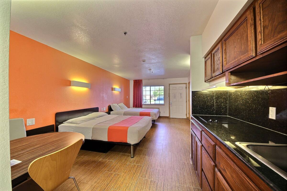 A hotel room with two beds, a wooden desk, a kitchenette, and orange walls.