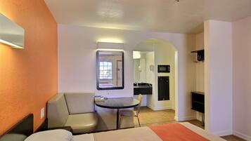 A hotel room with a bed, a desk, a chair, a small table, a mirror, and a kitchenette area.