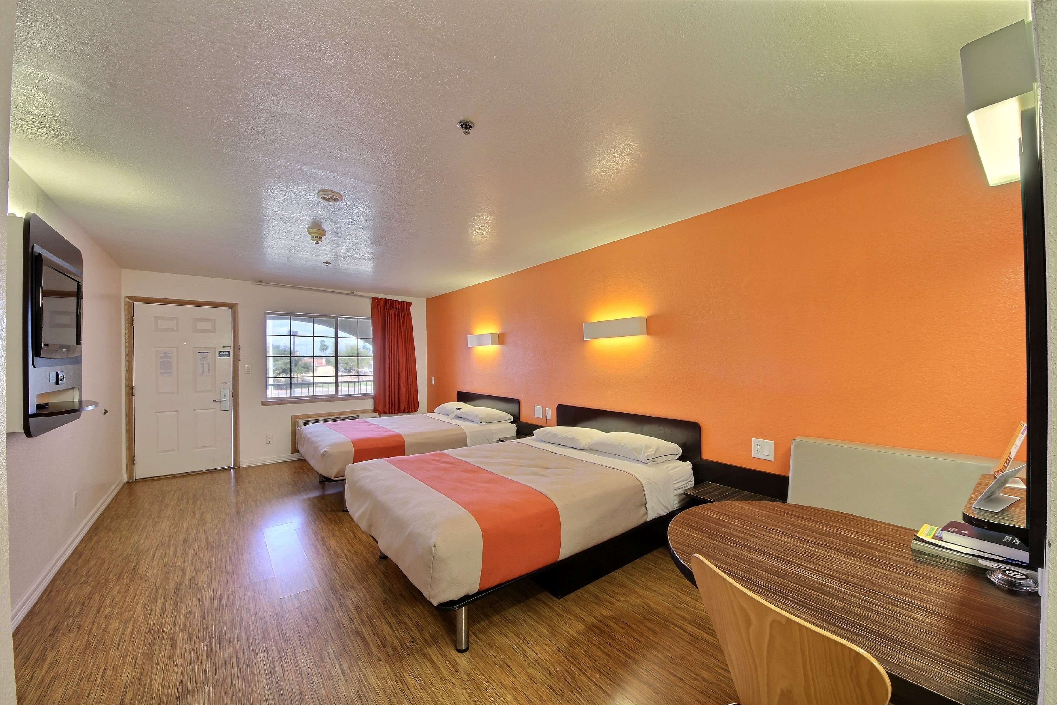 A hotel room with two beds, a wooden desk, and a window with orange curtains.