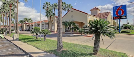 A motel with a clear view of the parking lot, palm trees, and a building with a red-tiled roof.