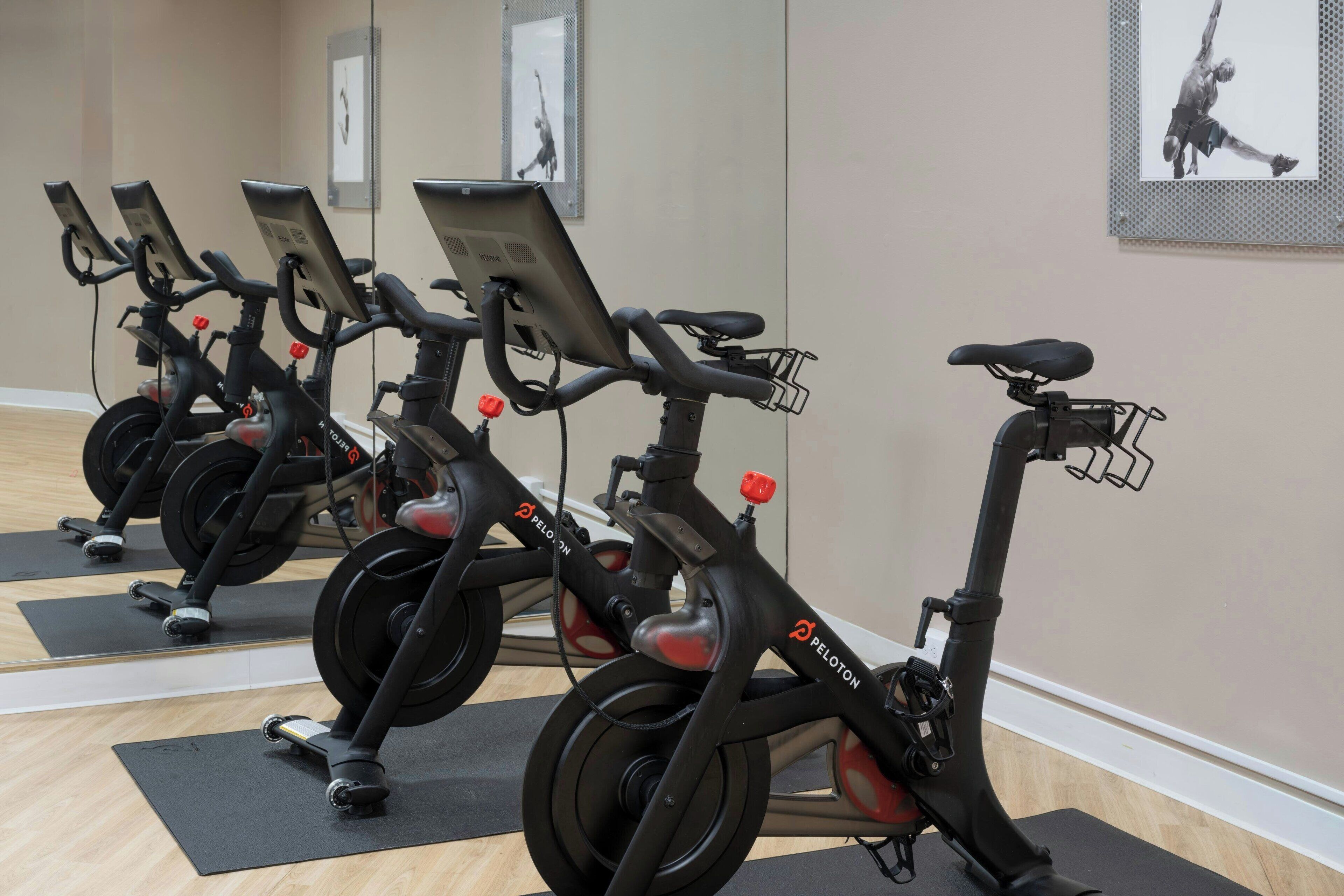 A row of stationary bikes in a gym with mirrors on the wall and exercise mats on the floor.