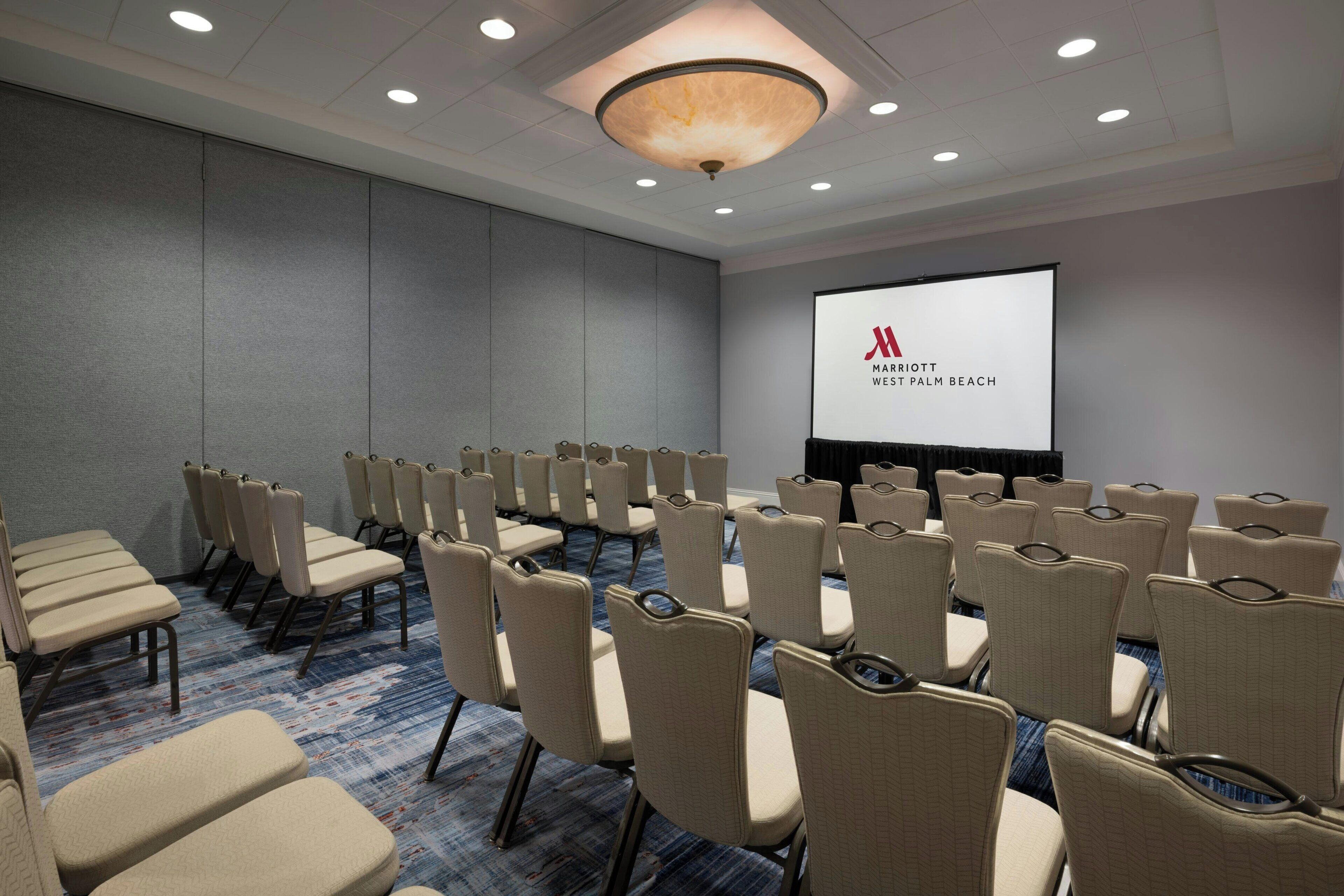A conference room with rows of beige chairs, a large screen, and a carpeted floor.