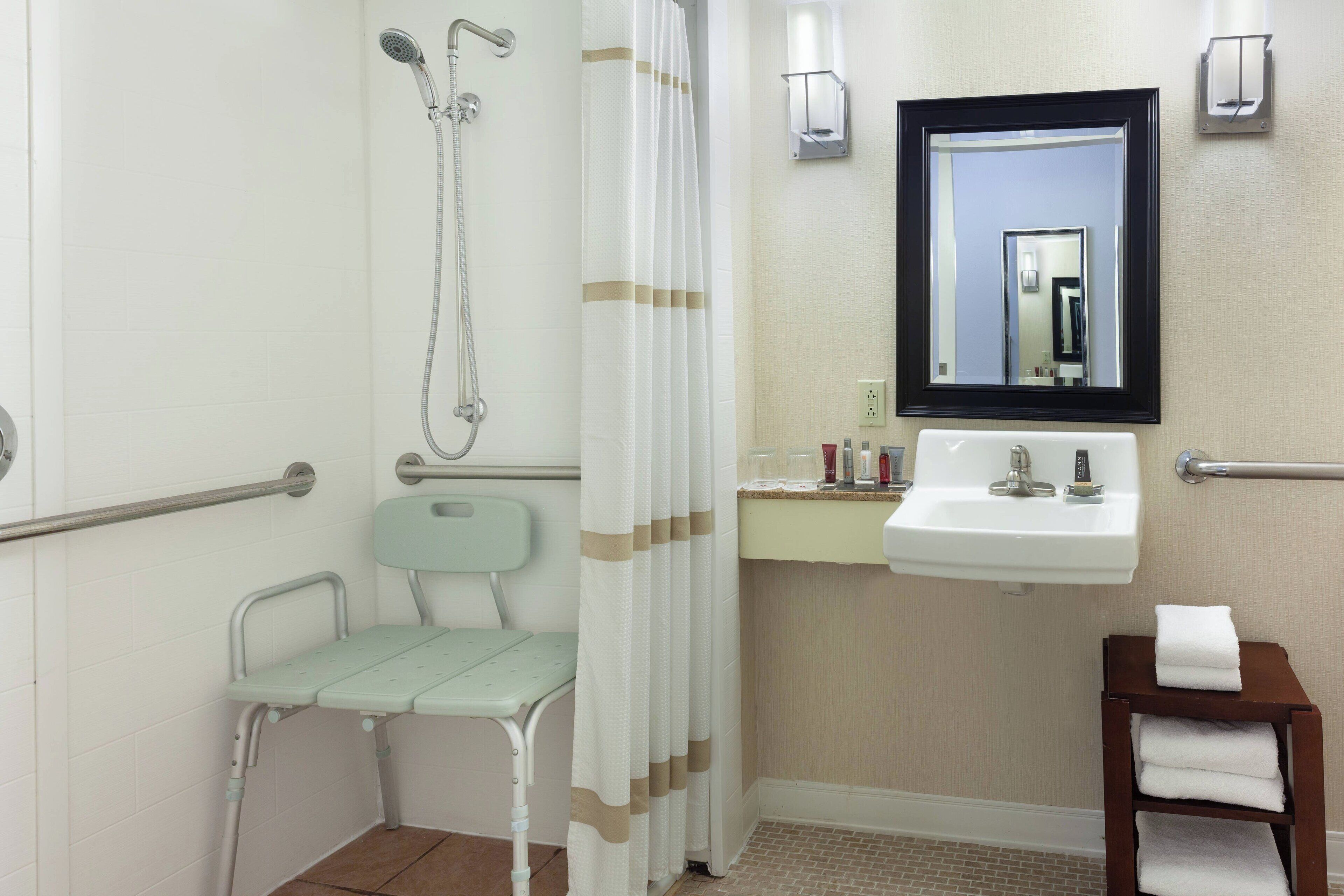 A bathroom with a shower, a sink, a mirror, a chair, and a small table with towels.