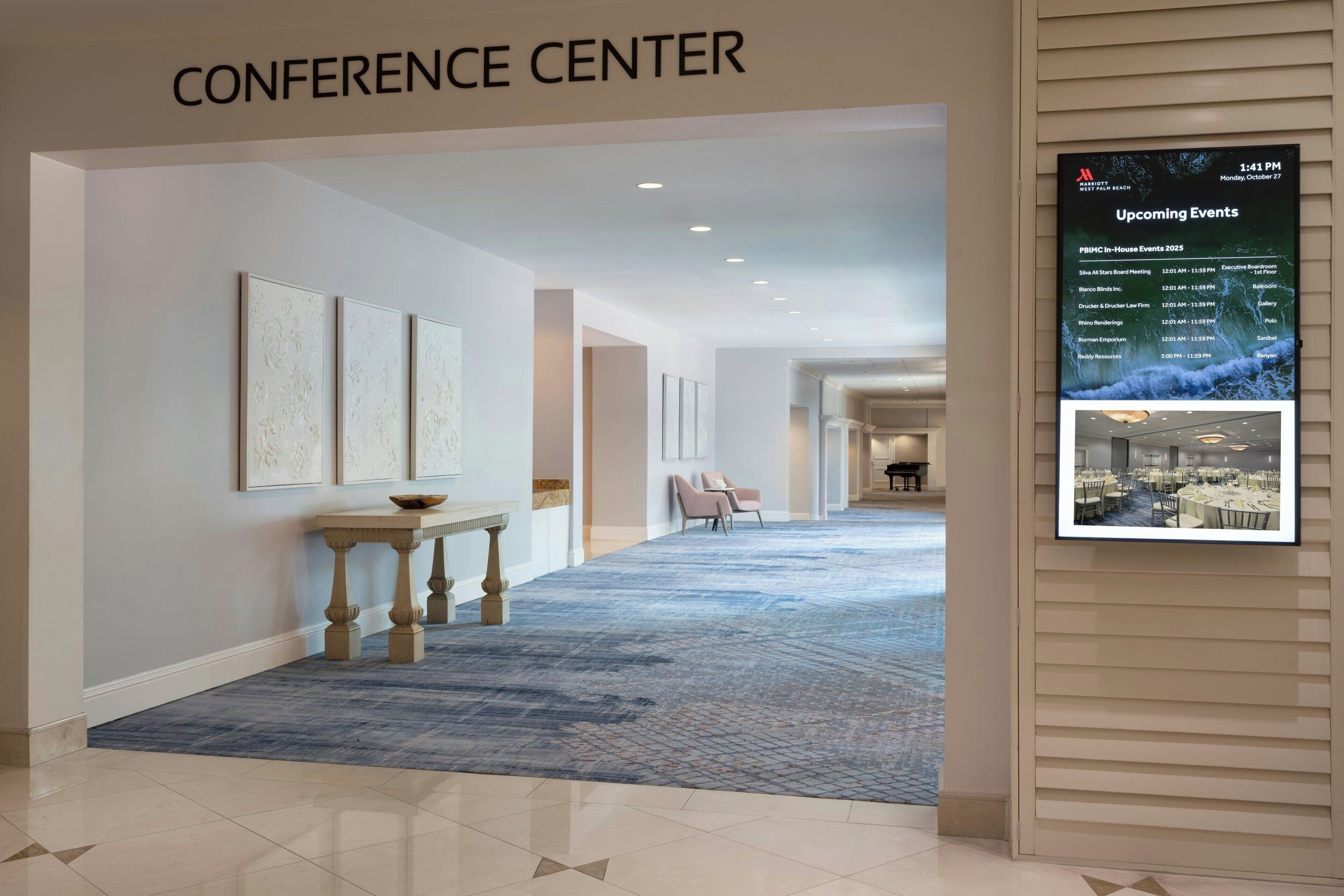 A conference center with a long hallway, framed artwork on the walls, a stone table, and a carpeted floor.