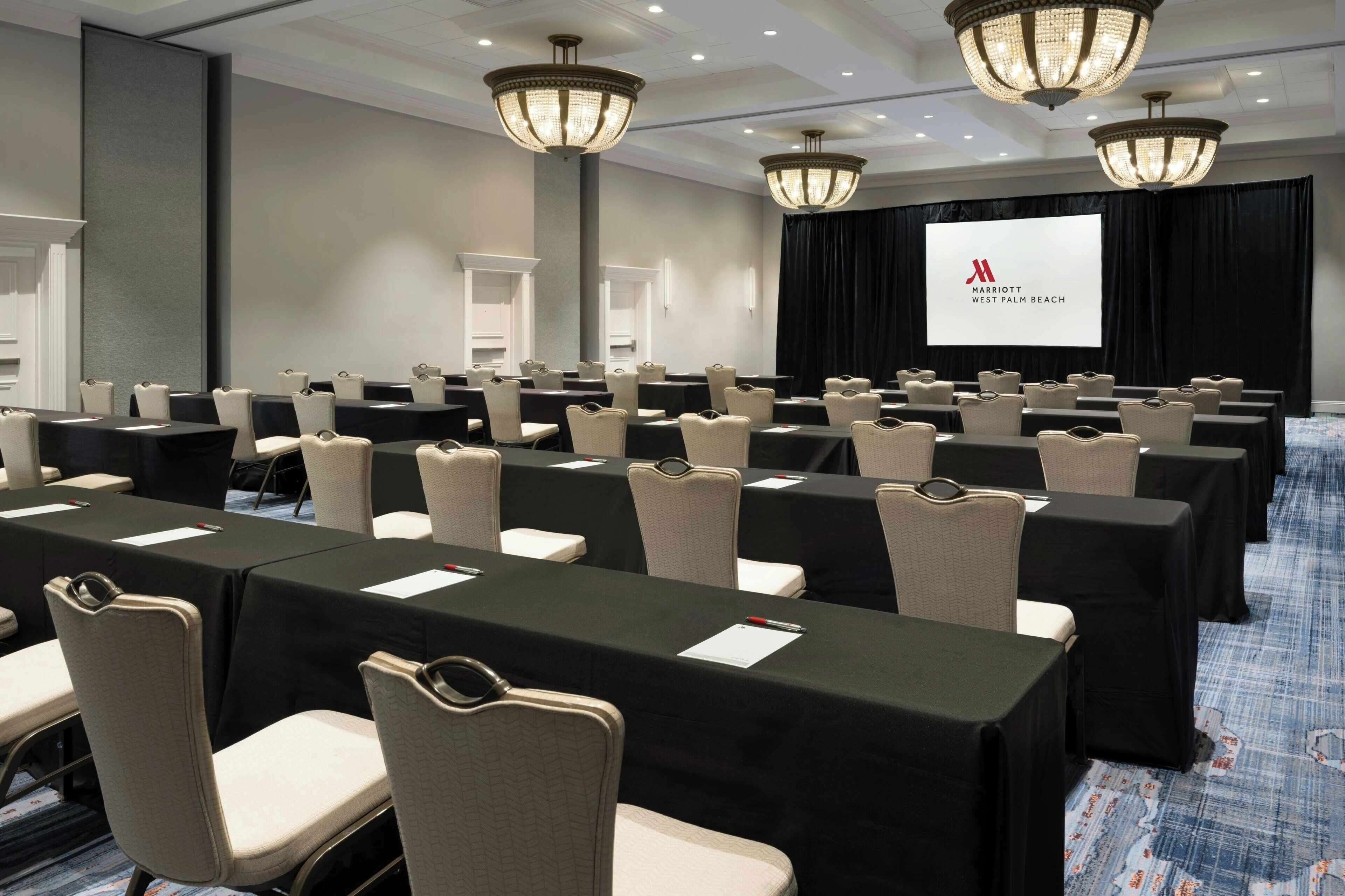 A conference room with rows of tables and chairs, a projector screen, and chandeliers.