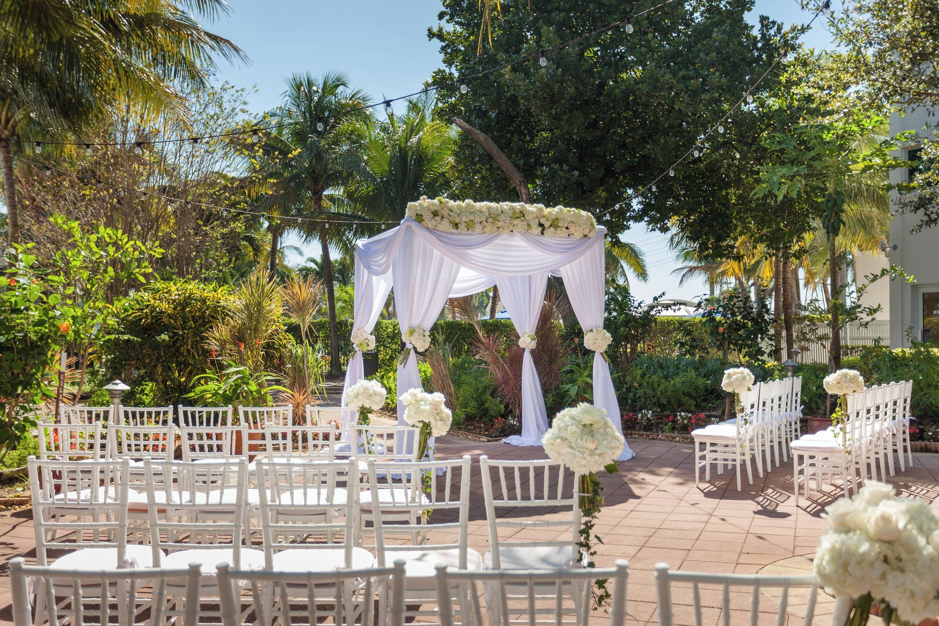 An outdoor wedding setup with white chairs and tables arranged in rows, a white canopy with floral decorations, and lush greenery in the background.