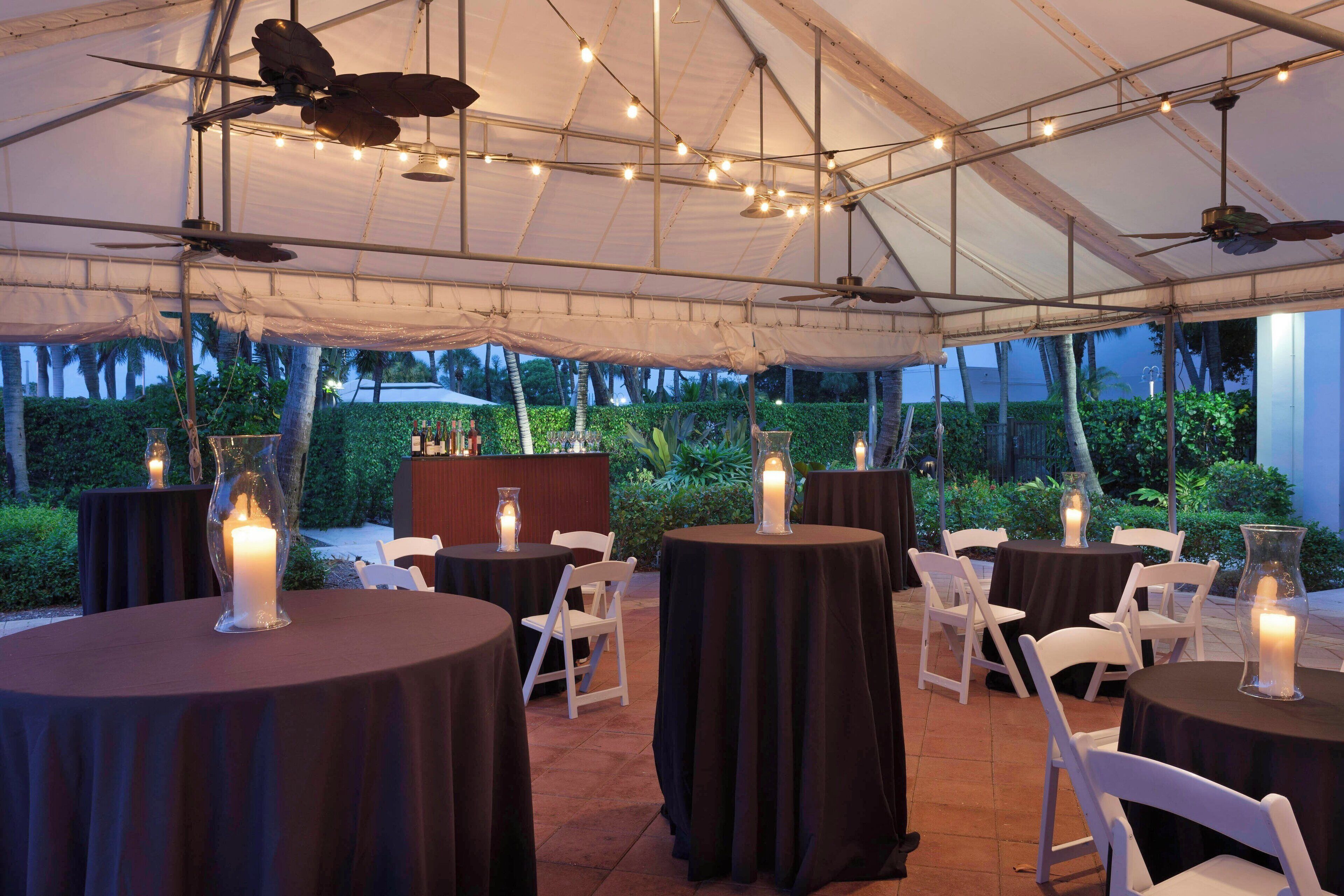 Un espacio para eventos al aire libre con mesas redondas cubiertas con telas oscuras, sillas blancas y velas.