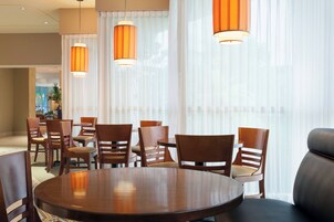 Un comedor con una mesa redonda y varias sillas, ventanas amplias con cortinas translúcidas y lámparas colgantes naranjas.