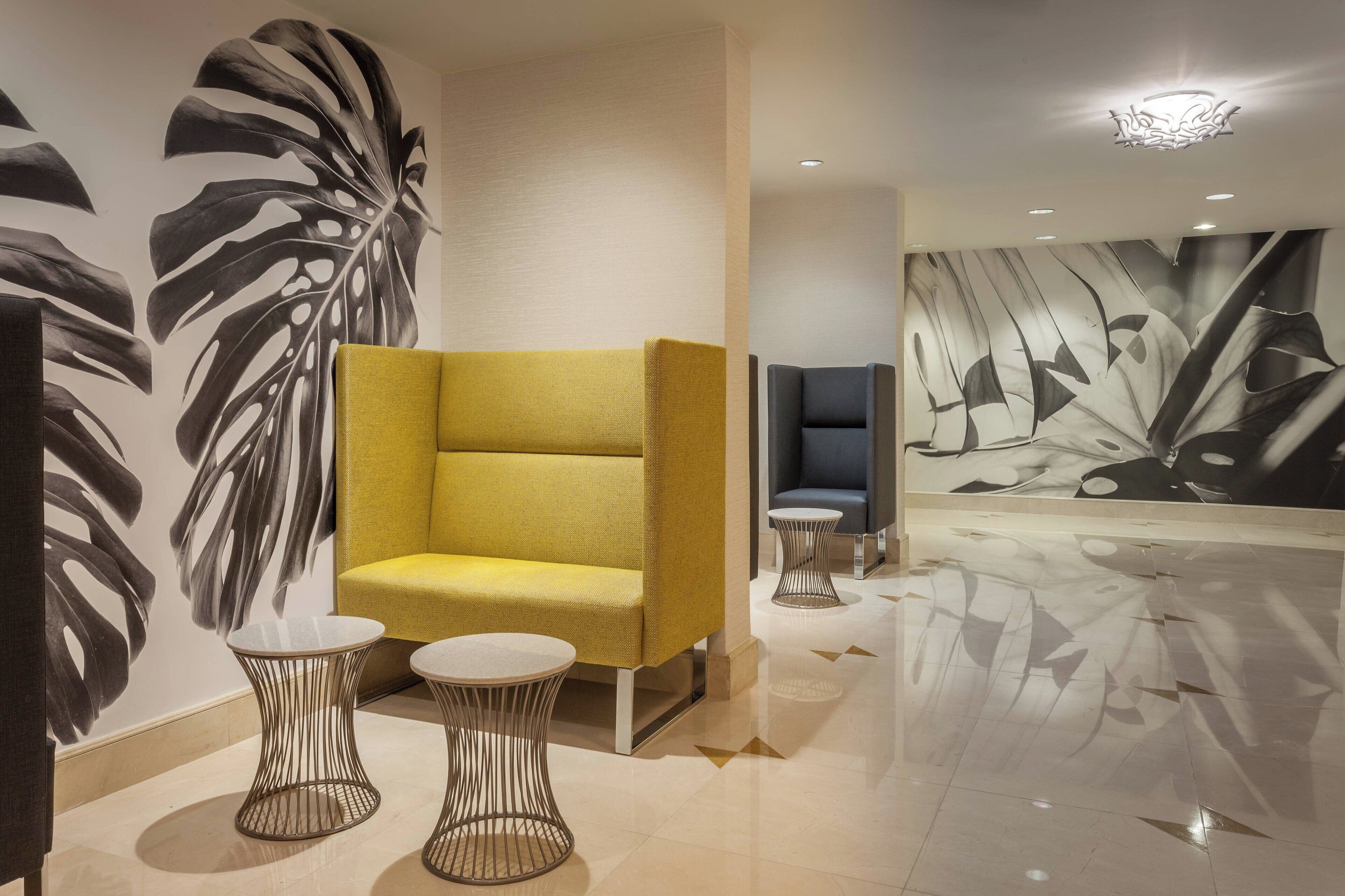 A modern lobby with a yellow sofa, two wireframe tables, and a large abstract mural on the wall.