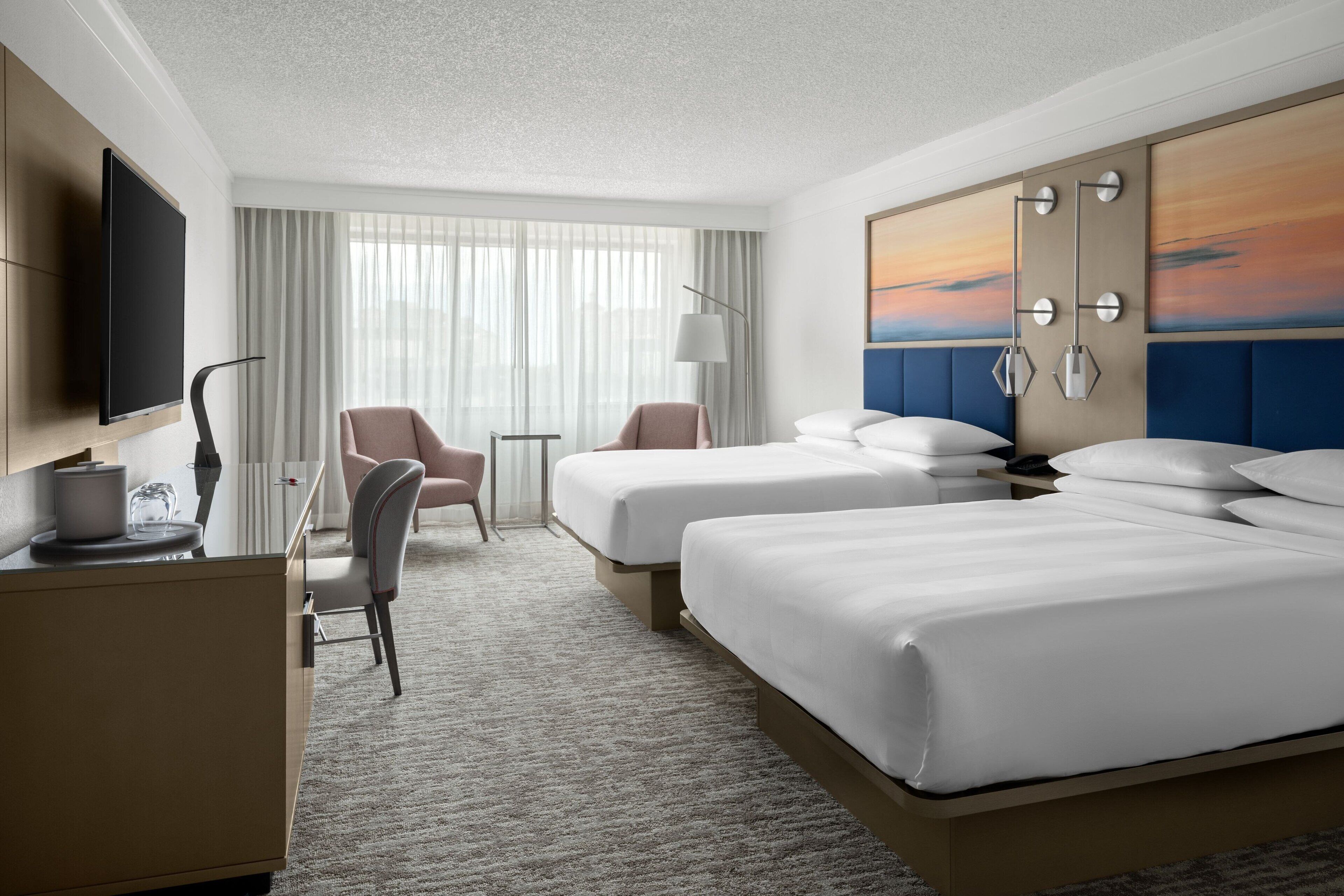 Quarto de hotel com duas camas, uma escrivaninha, uma cadeira, uma televisão e um quadro na parede.