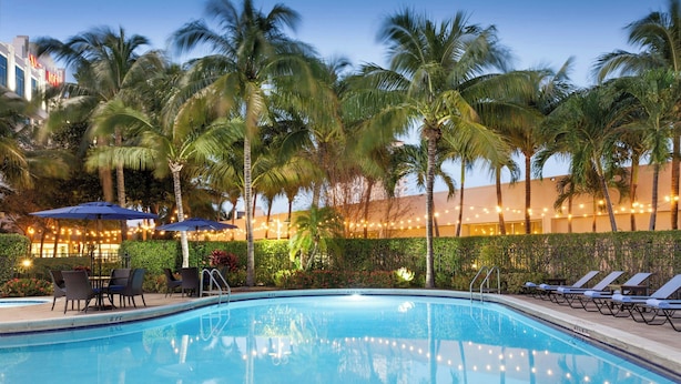 A swimming pool with lounge chairs and umbrellas, surrounded by palm trees and string lights.