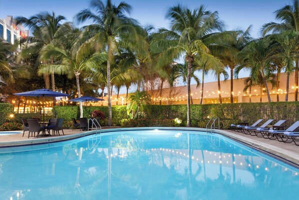 A swimming pool with lounge chairs and umbrellas, surrounded by palm trees and string lights.