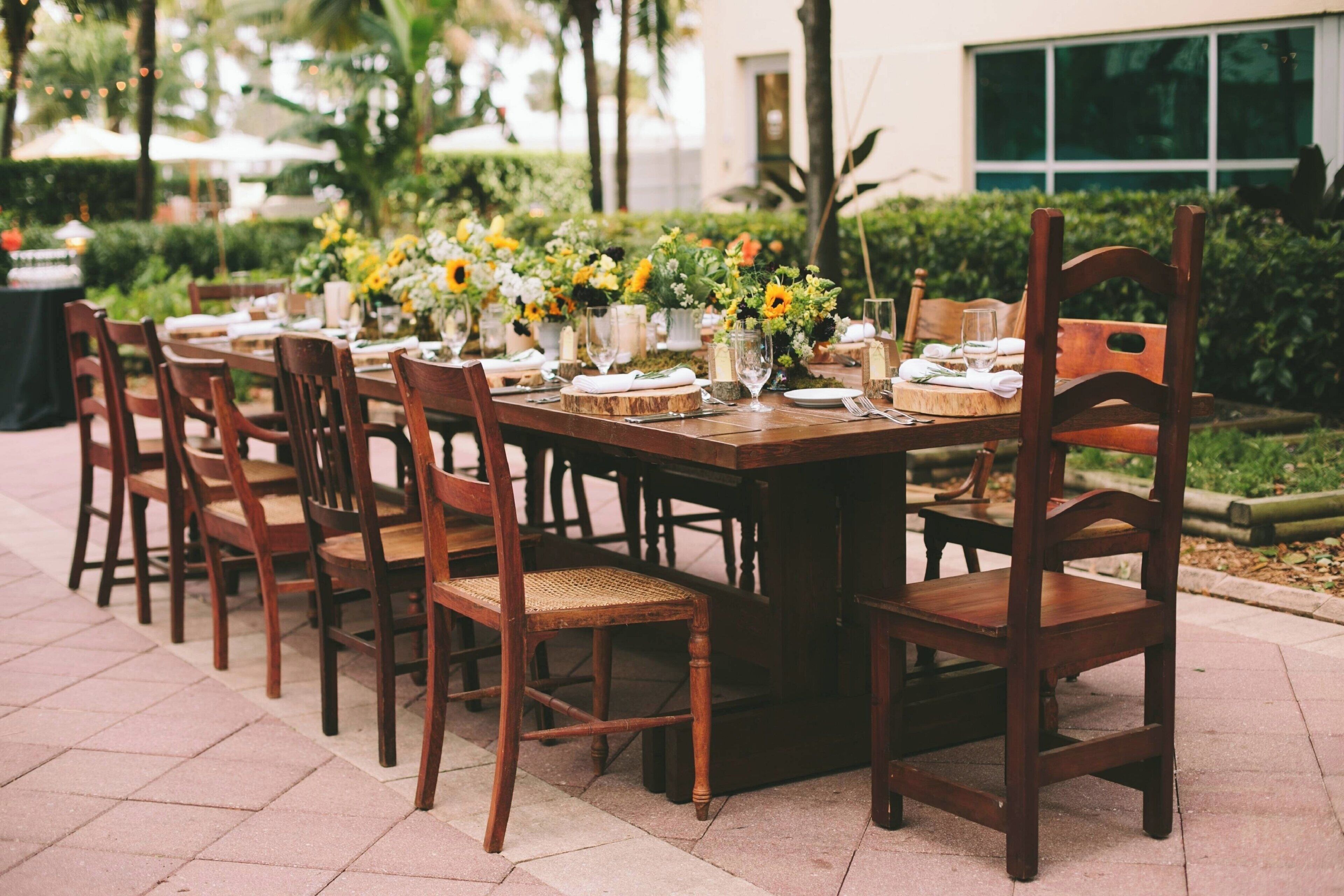 An outdoor dining area with wooden tables and chairs, surrounded by greenery and a building in the background.